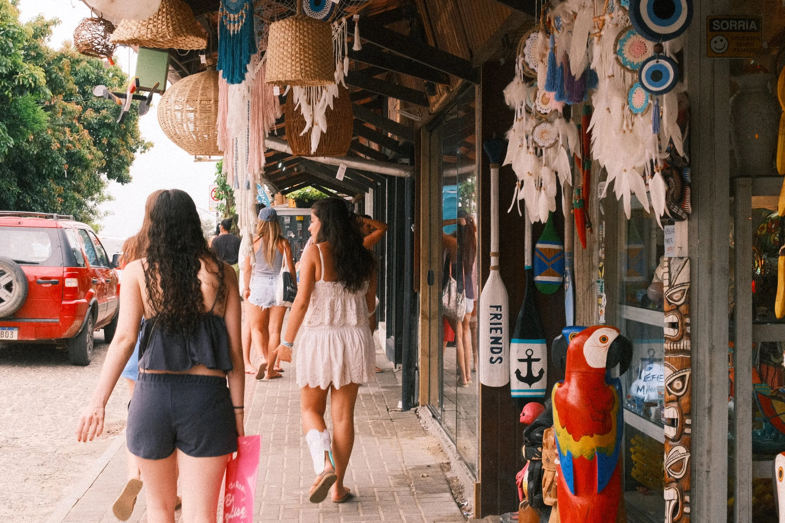 People walking on a sidewalk along a shop with colorful handmade crafts, such as dreamcatchers, painted parrots, and wooden masks, in a tropical setting.