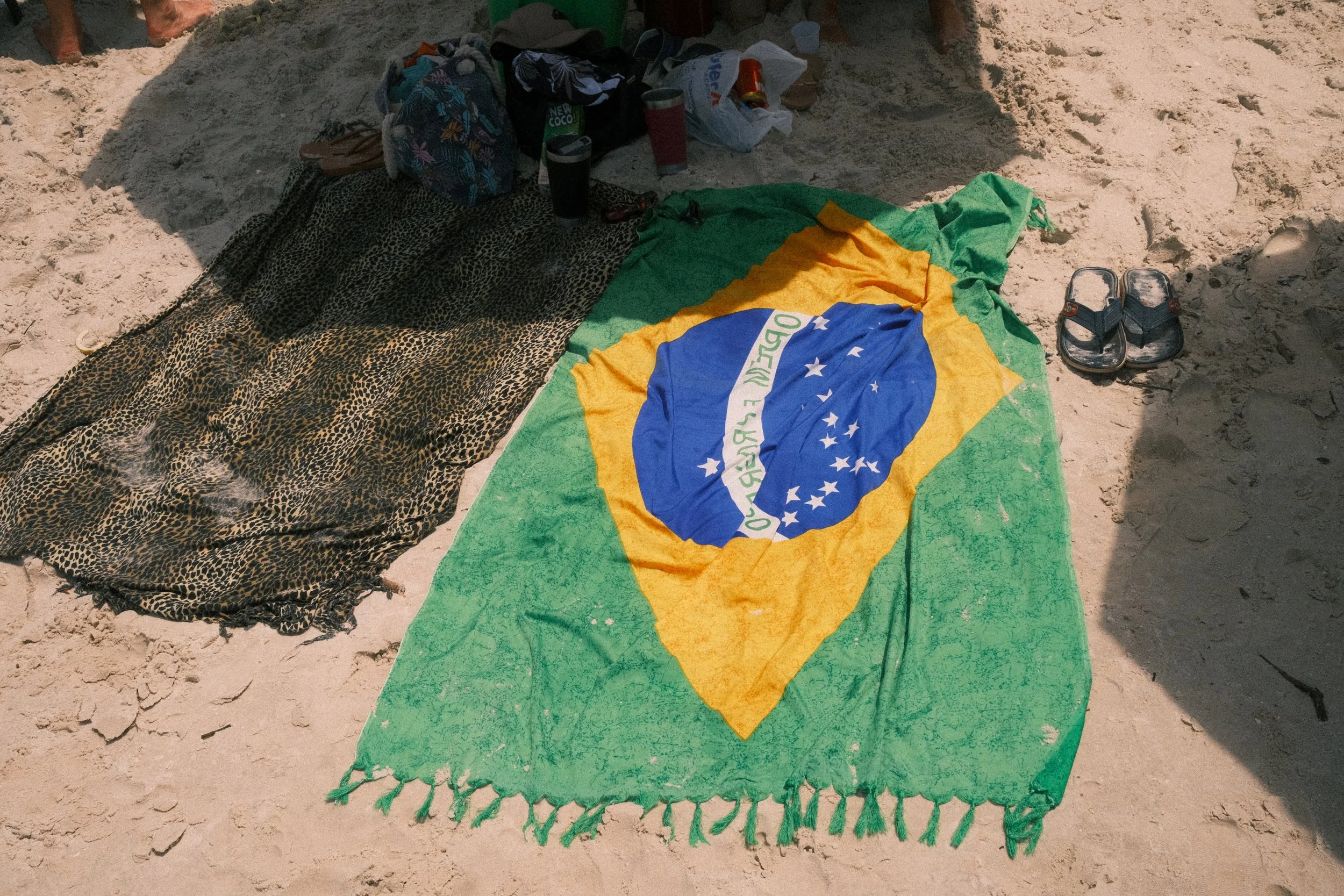 Beach scene with towels and belongings, including a Brazilian flag towel on the sand, shoes, and containers.