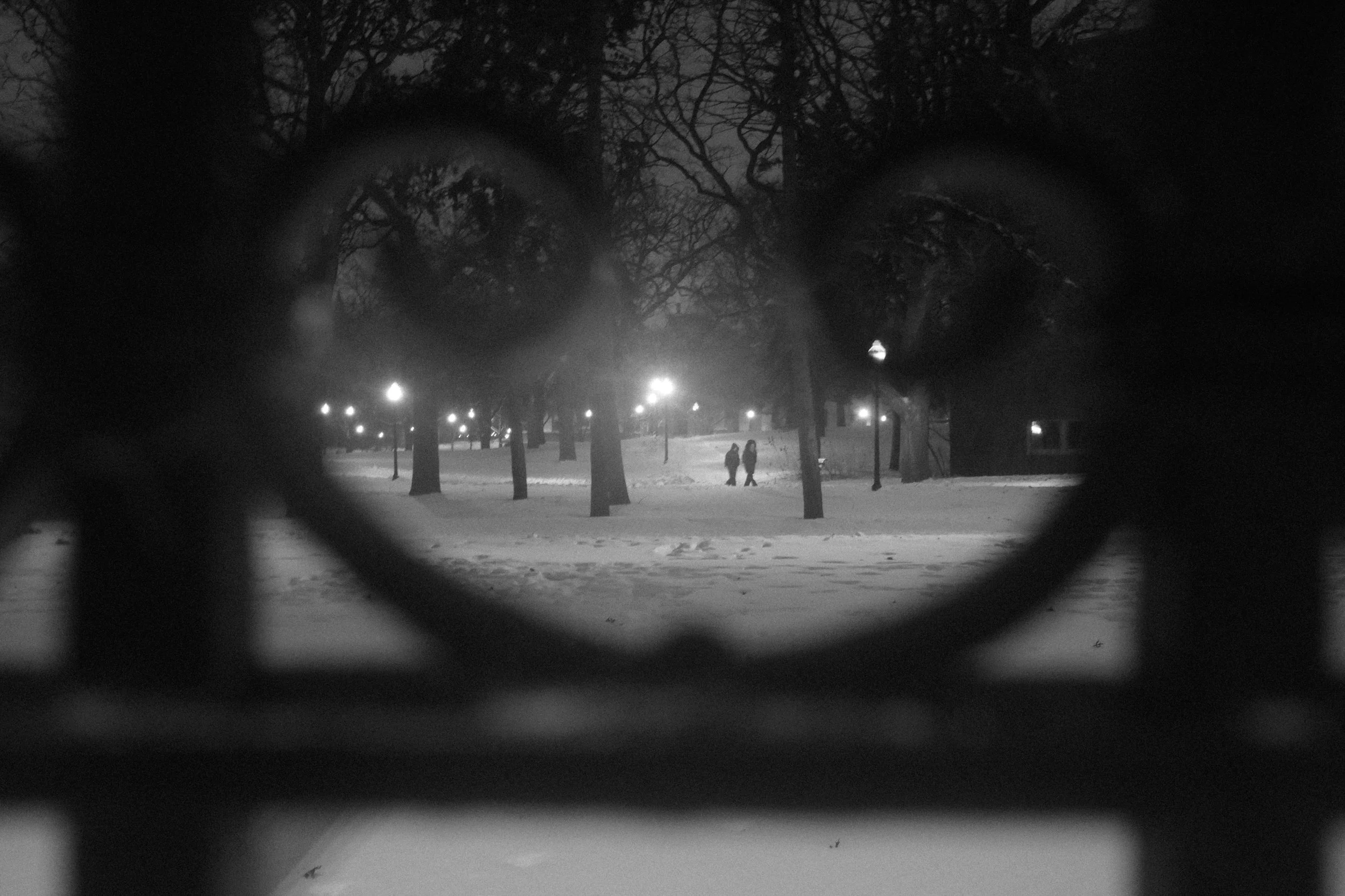 Nighttime park scene seen through a pair of glasses on a snow-covered landscape with people walking and streetlights illuminating the area.