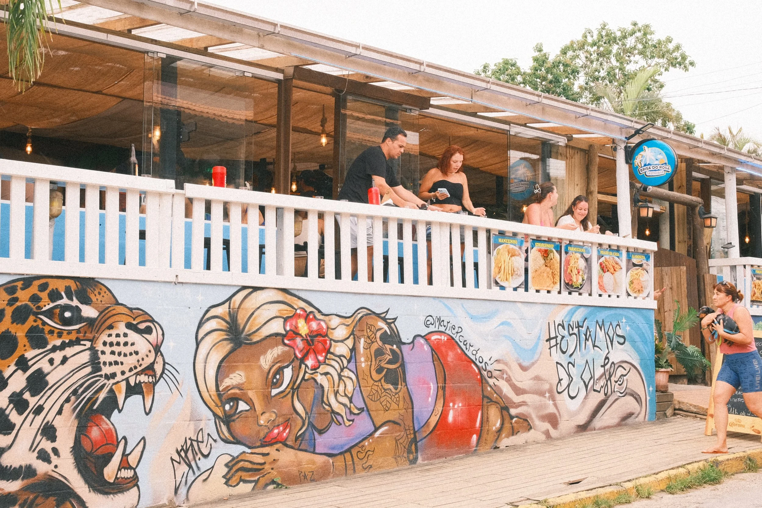 People dining on an outdoor balcony with a mural of a woman with blonde hair, tattoos, a red dress, and a red hibiscus in her hair. The mural is painted on a wall with a drawing of a jaguar.