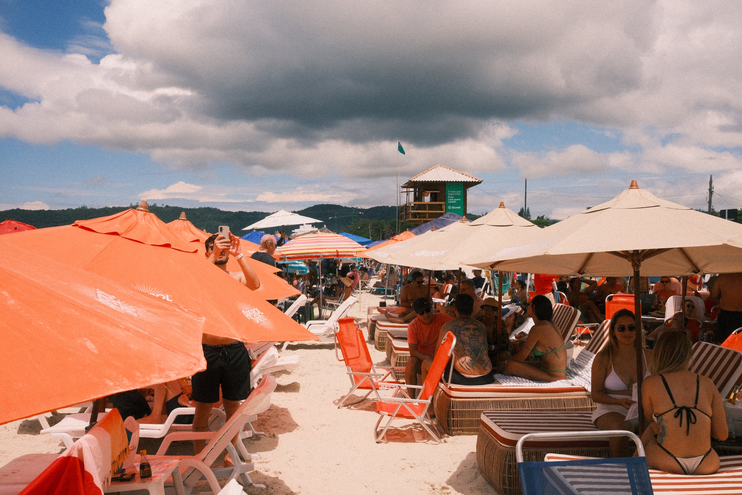 A crowded beach scene with many orange and white umbrellas, lounge chairs, and people relaxing and socializing under partly cloudy skies.