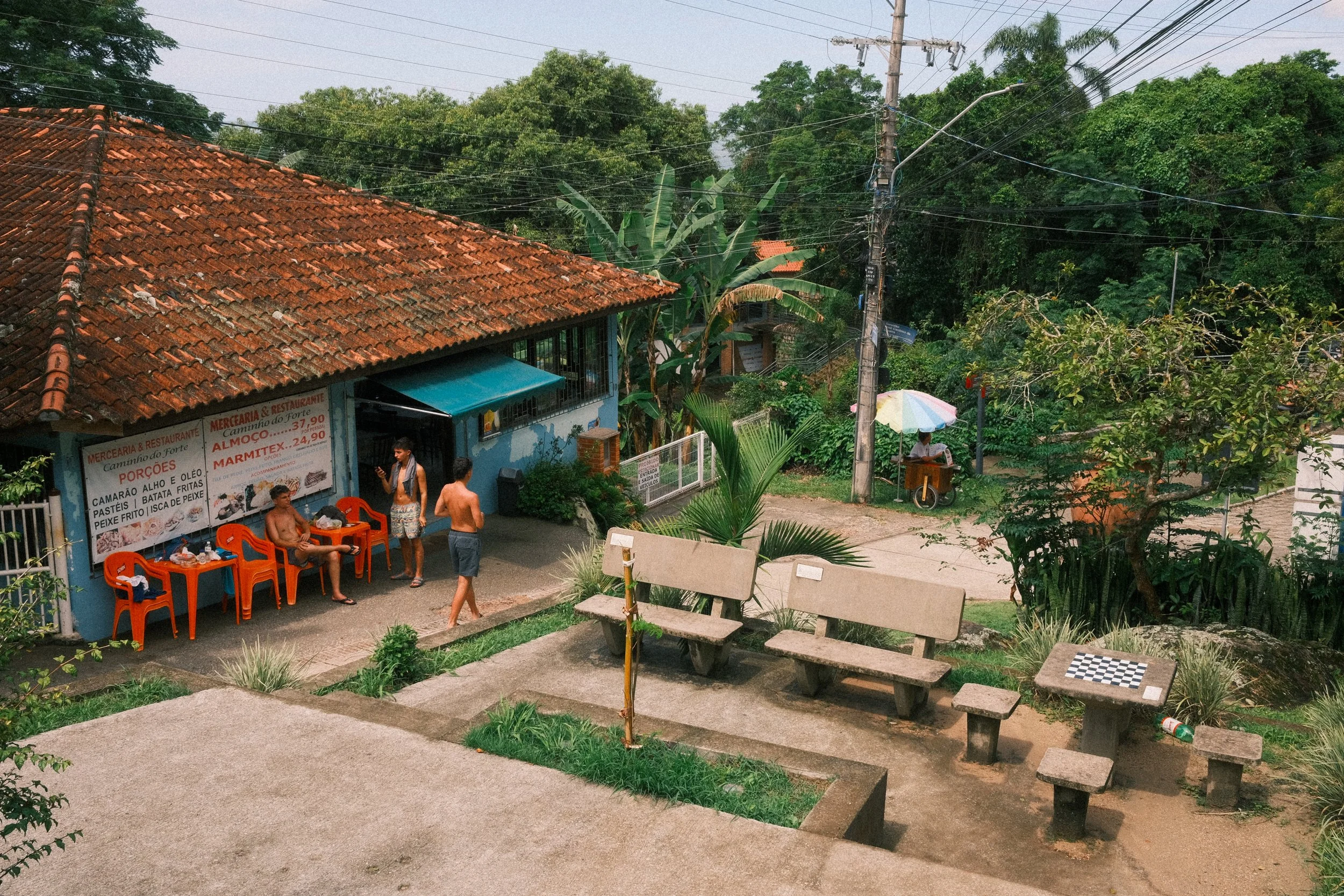 A small street-side eatery with a tiled roof, blue walls, and four orange chairs outside, one of which a man is sitting on. Three men are near the entrance, and a person under a colorful umbrella is beside a bicycle cart. In the foreground, there are concrete benches and a table with a checkerboard on it, surrounded by greenery.