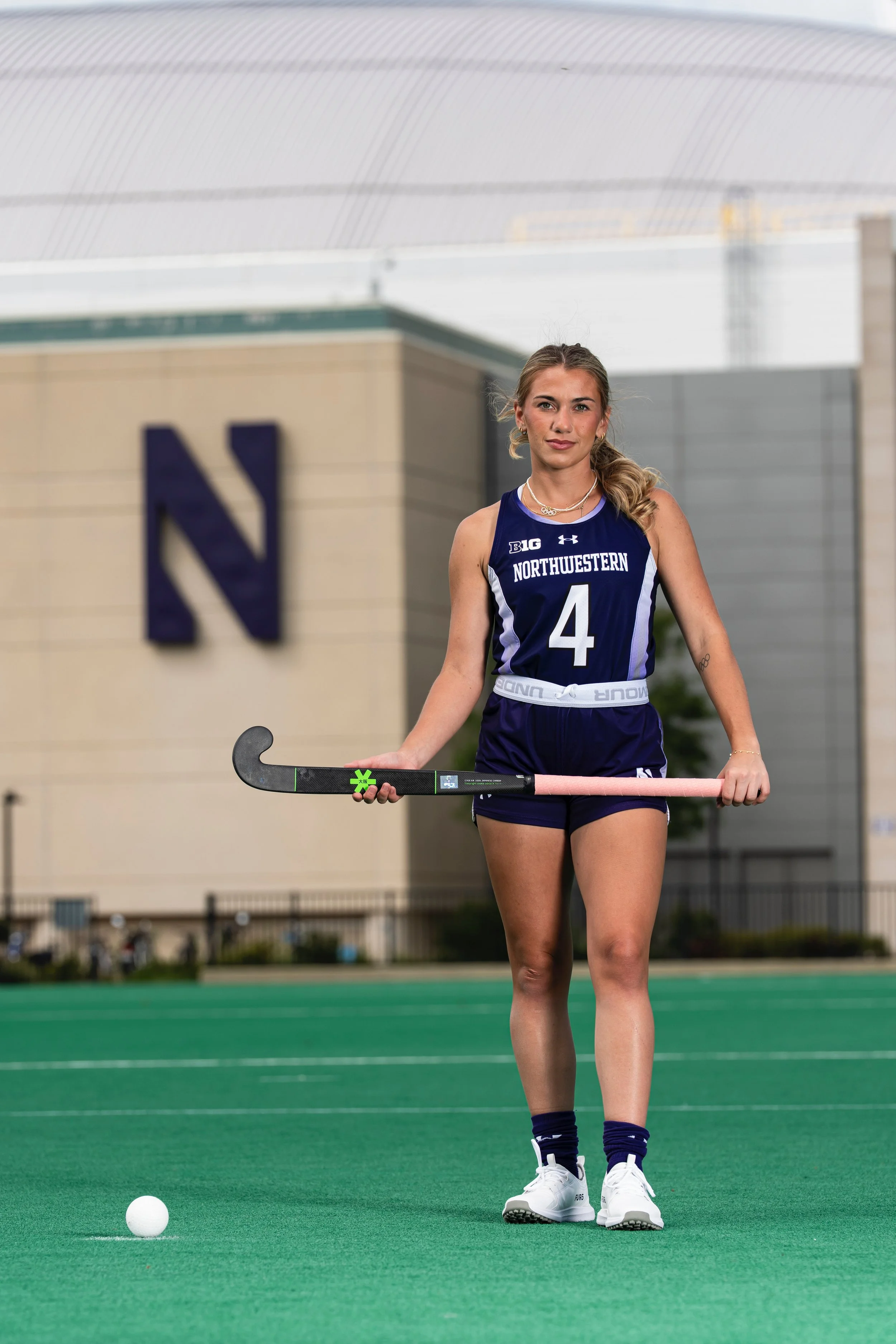 A young female field hockey player wearing a navy Northwestern uniform with the number 4, holding a pink stick, standing on a green field with a white ball nearby, in front of a large building with a prominent N logo.