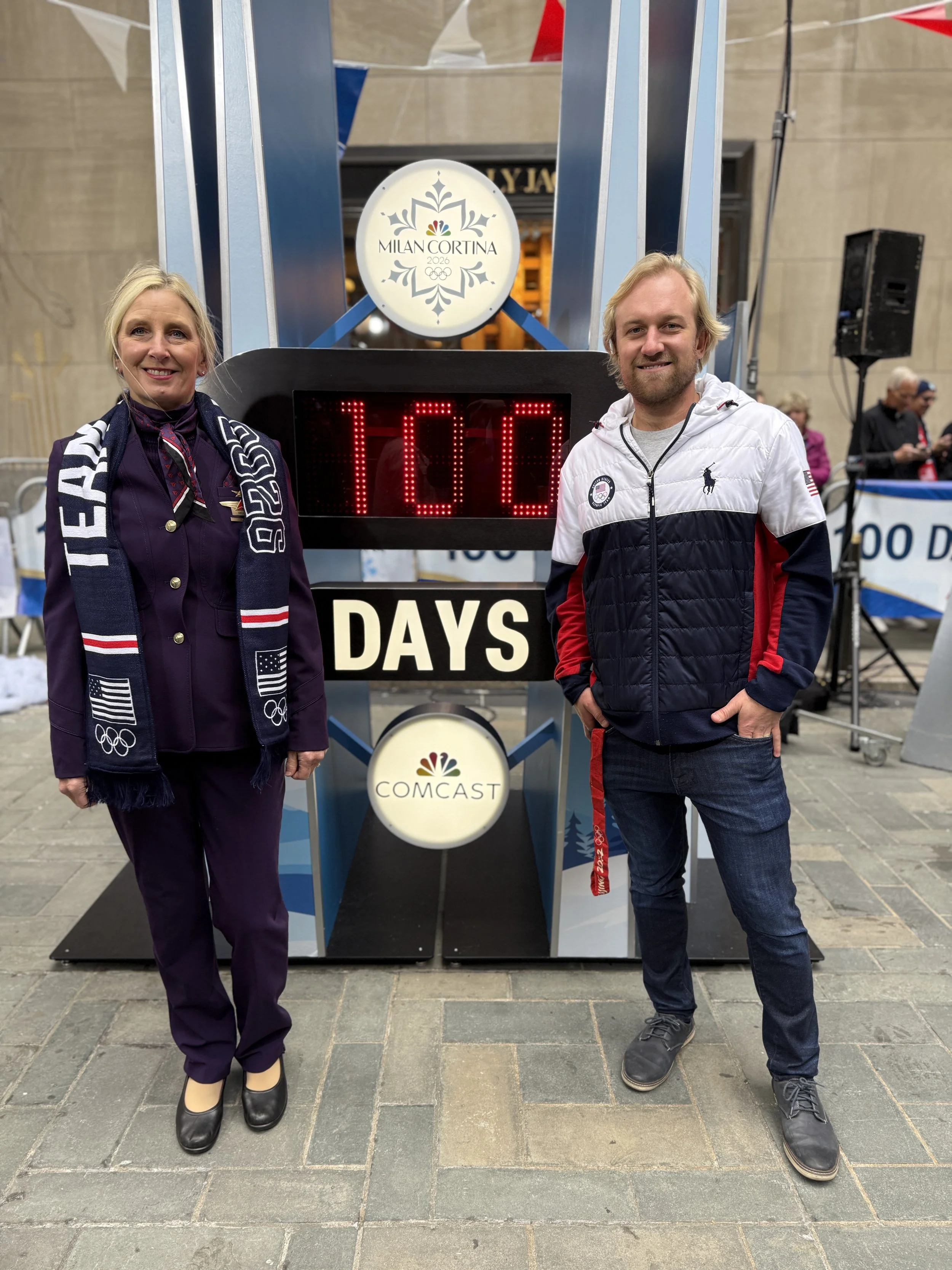 Two people standing in front of a countdown clock that shows 100 days, with signs for the Milano Cortina 2026 Winter Olympics and NBC Comcast. The woman on the left is wearing a dark outfit with a scarf, and the man on the right is wearing a white, red, and navy jacket with jeans.