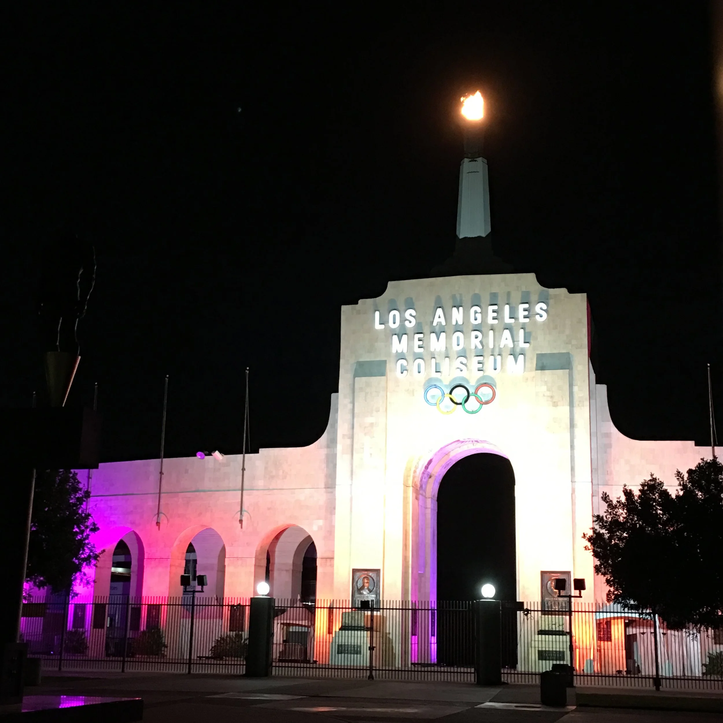 Night view of the Los Angeles Memorial Coliseum entrance with the Olympic rings and a flaming torch on top, illuminated with colorful lights.