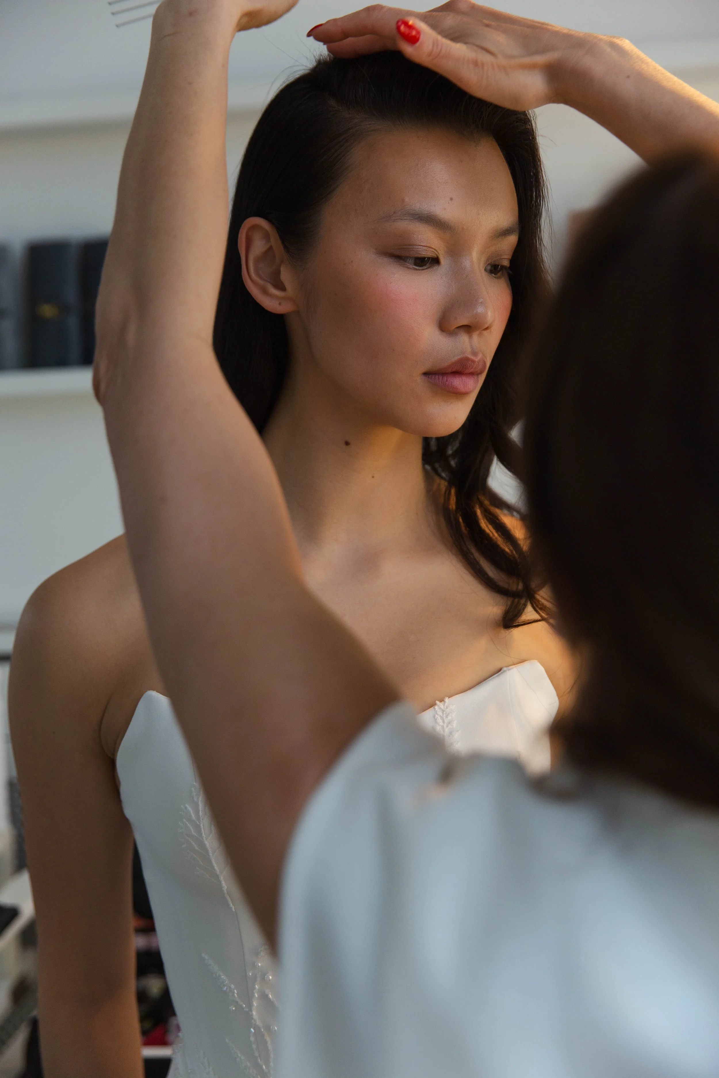 A woman with dark hair is having her hair styled by another person with red-painted nails; she is dressed in a strapless white dress and appears to be in a dressing room with shelves of black books or boxes in the background.