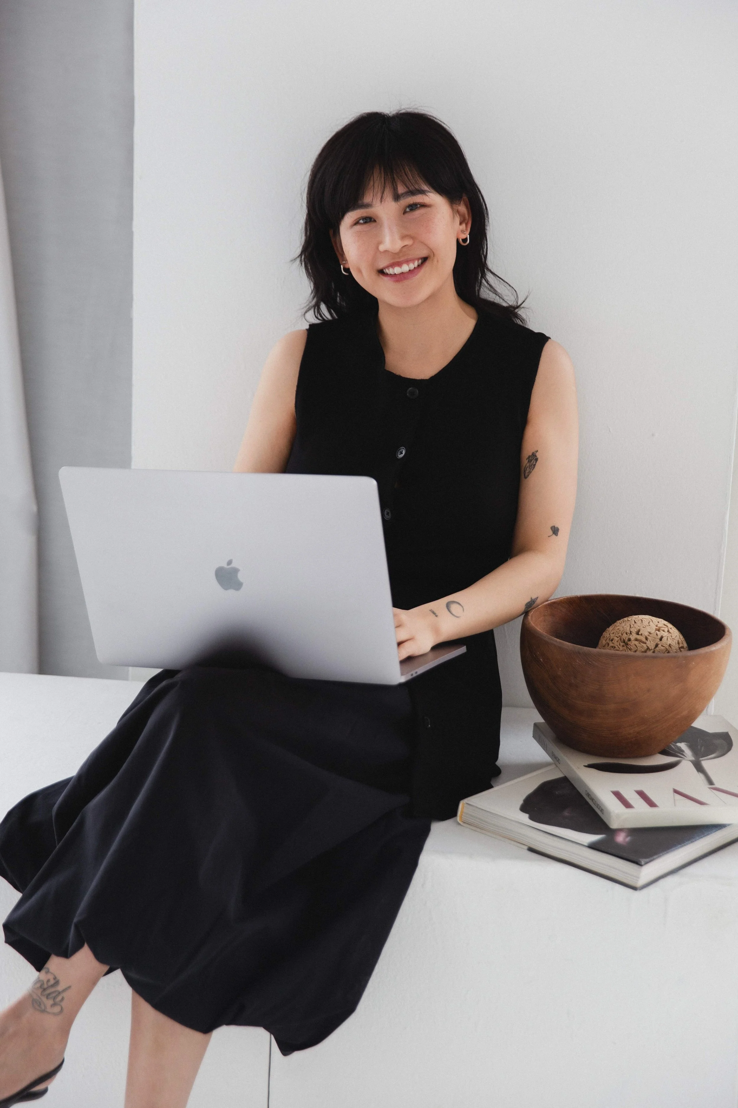 Lächelnde junge Frau mit Tattoos sitzt bei einem weißen Tisch mit einem MacBook, einem großen Holschale mit Dekor und mehreren Büchern.