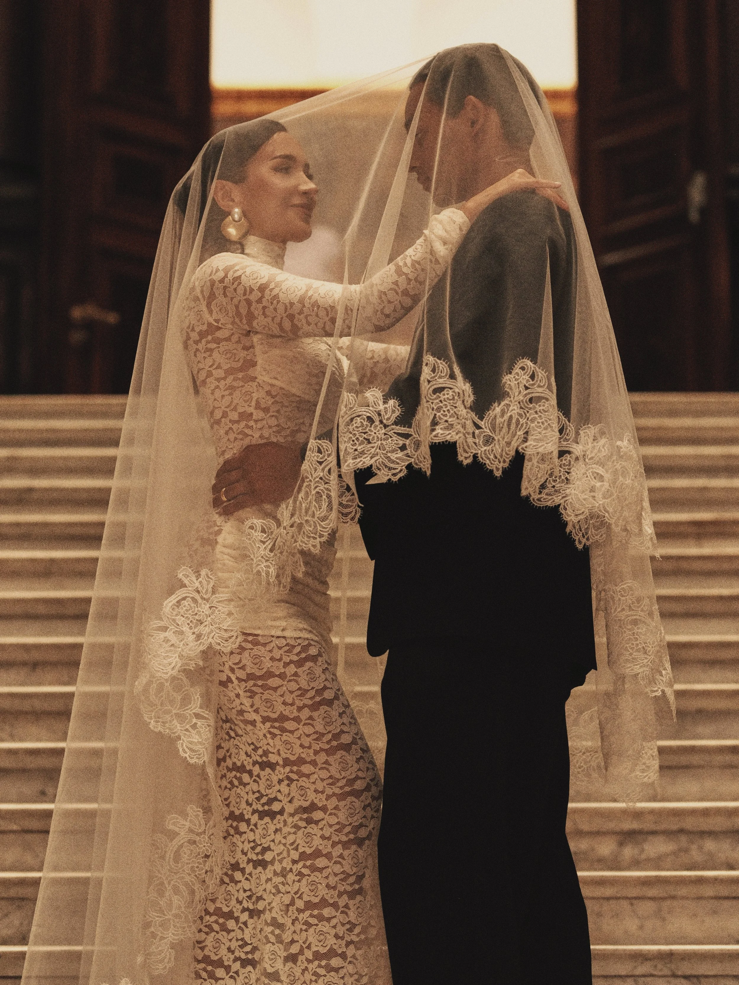 Ein Brautpaar steht im elegant gedeckten Innenraum, die Braut trägt ein Spitzenkleid und eine Hochzeitsschleier, während der Bräutigam einen dunklen Anzug trägt. Sie stehen auf einer Treppe und teilen einen intimen Moment.