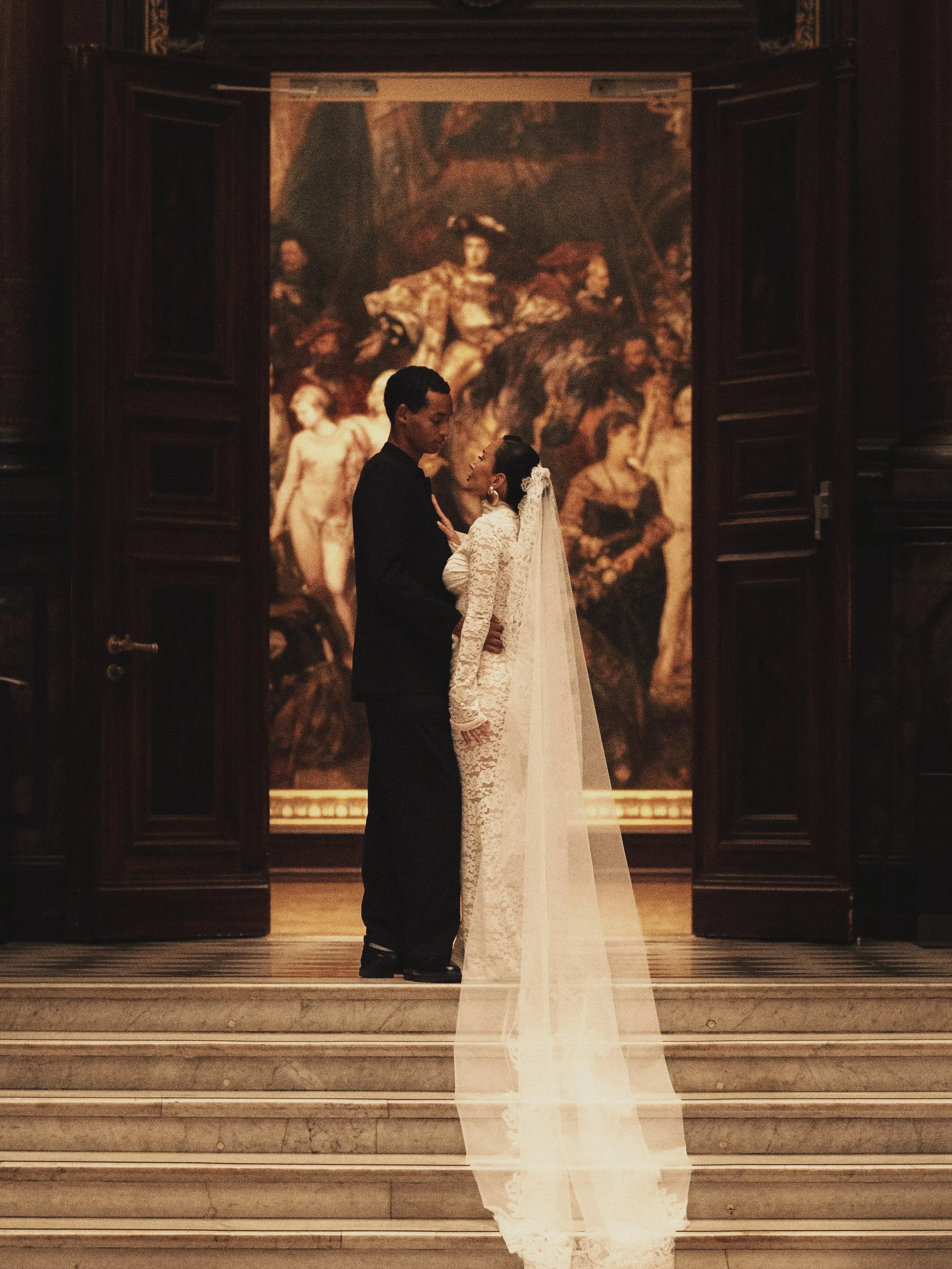 Ein Paar in formeller Kleidung, ein Mann in einem schwarzen Anzug und eine Frau in einem weißen Hochzeitskleid, stehen sich in einem Museum oder Kunsthalle in einer Nähe, umgeben von Gemälden und Holzrahmen.
