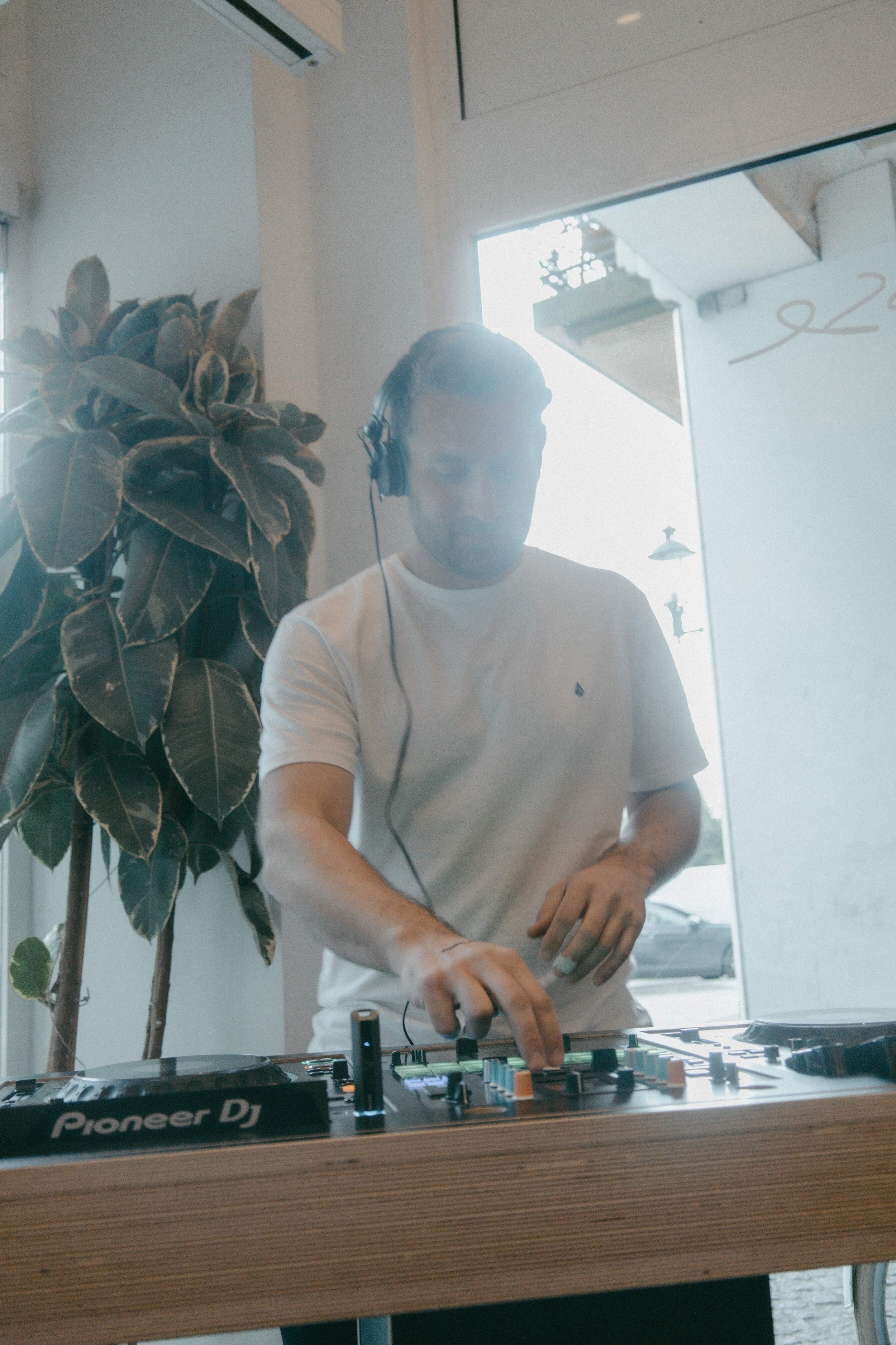 Ein junger Mann mit Kopfhörern, der an einem DJ-Mischer arbeitet, mit einem Pflanzen im Hintergrund und einem Spiegel an der Wand.