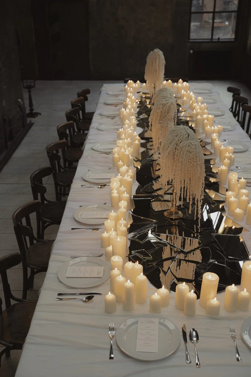 Ein lange gedeckter Tisch mit weißen Kerzen, weißen Tellern, Besteck und eleganten Tischdekorationen, darunter weiße Blumenarrangements auf schwarzen Spiegelplatten, in einem rustikalen Raum mit Fenstern im Hintergrund.