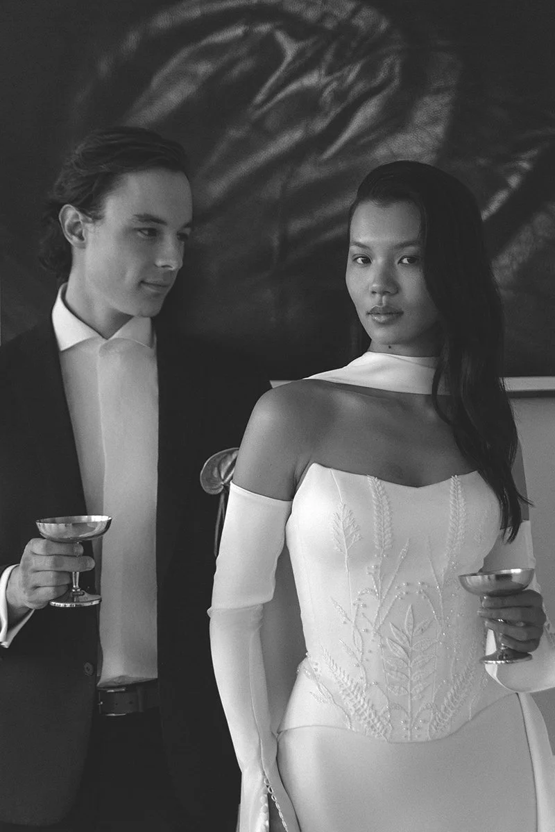 Ein schwarz-weiß Foto von einem Mann in einem Anzug und einer Frau in einem eleganten Kleid, die Getränke in Hand halten, vor einer künstlerischen Wanddekoration.