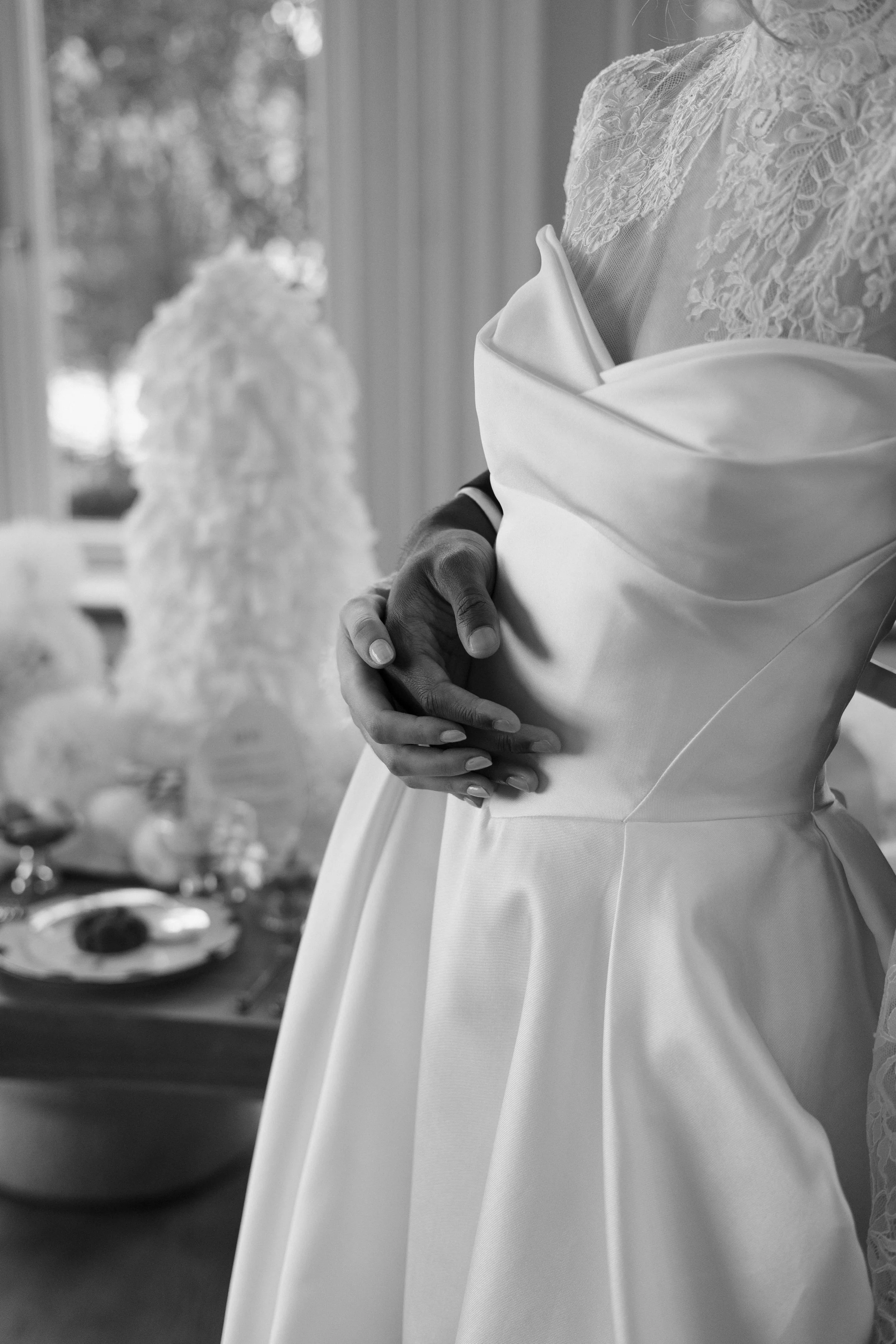 Nahaufnahme einer Person in einem weißen Hochzeitskleid, die die Arme um die Taille legt. Das Kleid hat einen satinierten Stoff mit Falten und Spitze am oberen Rand. Im Hintergrund ist ein Tisch mit einer Hochzeitstorte und Dekorationen zu sehen.