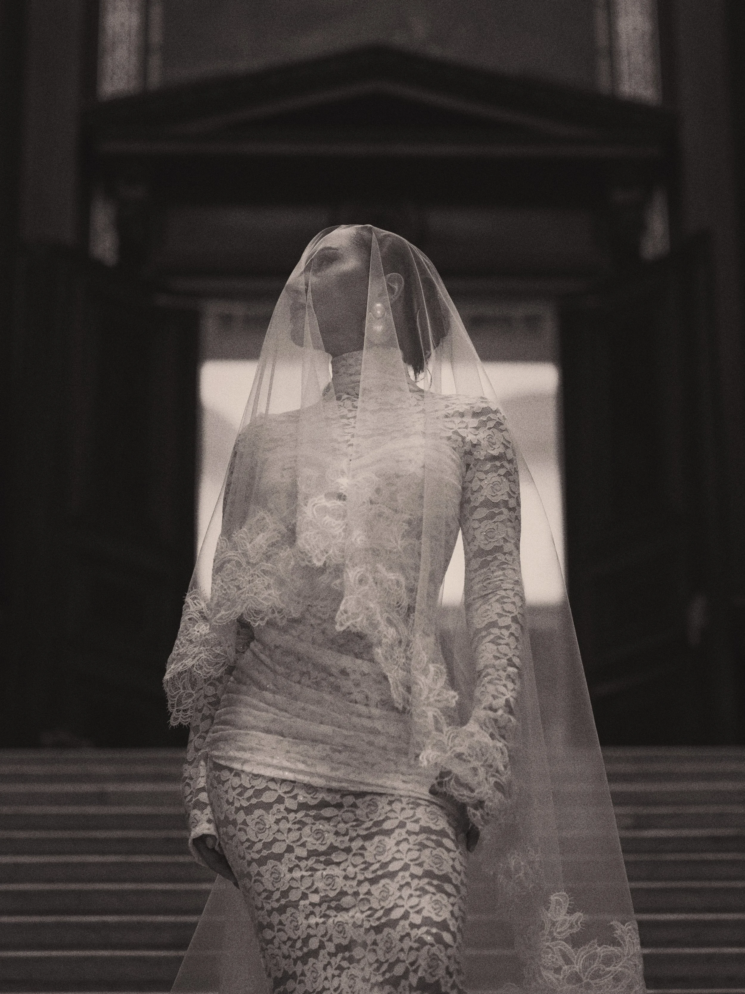 Eine Frau trägt ein hochzeitskleid mit Spitzenmuster und einen Schleier auf dem Kopf, steht auf einer Treppe im Gebäude.