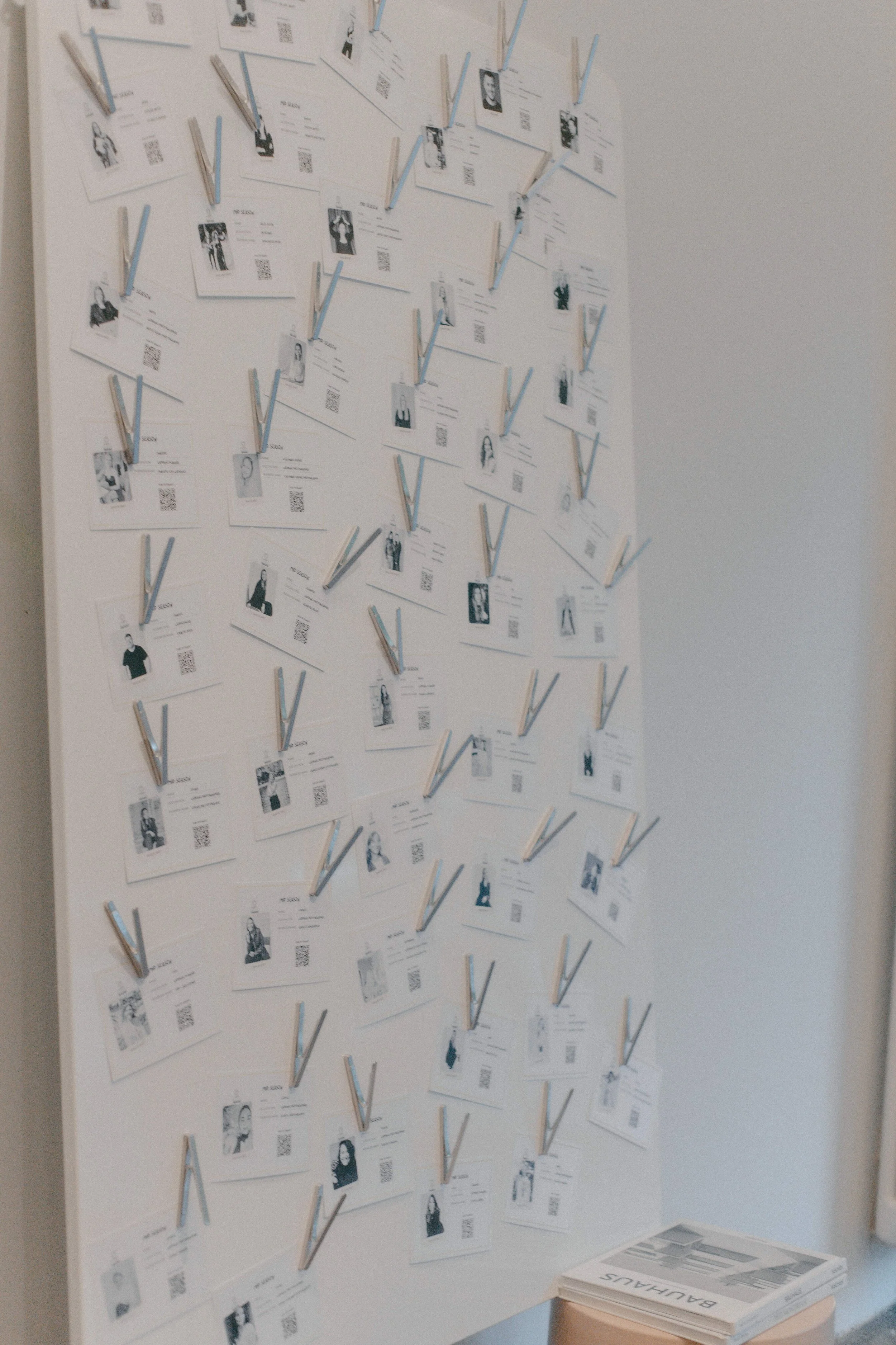 Ein Whiteboard ist mit mehreren Karten bedeckt, die Fotos, QR-Codes und Text enthalten, und sind durch weiße und blaue Wäscheklammern angebracht. Es gibt einen Stapel Broschüren mit der Aufschrift 'BAUHAUS' auf einem kleinen Tisch daneben.