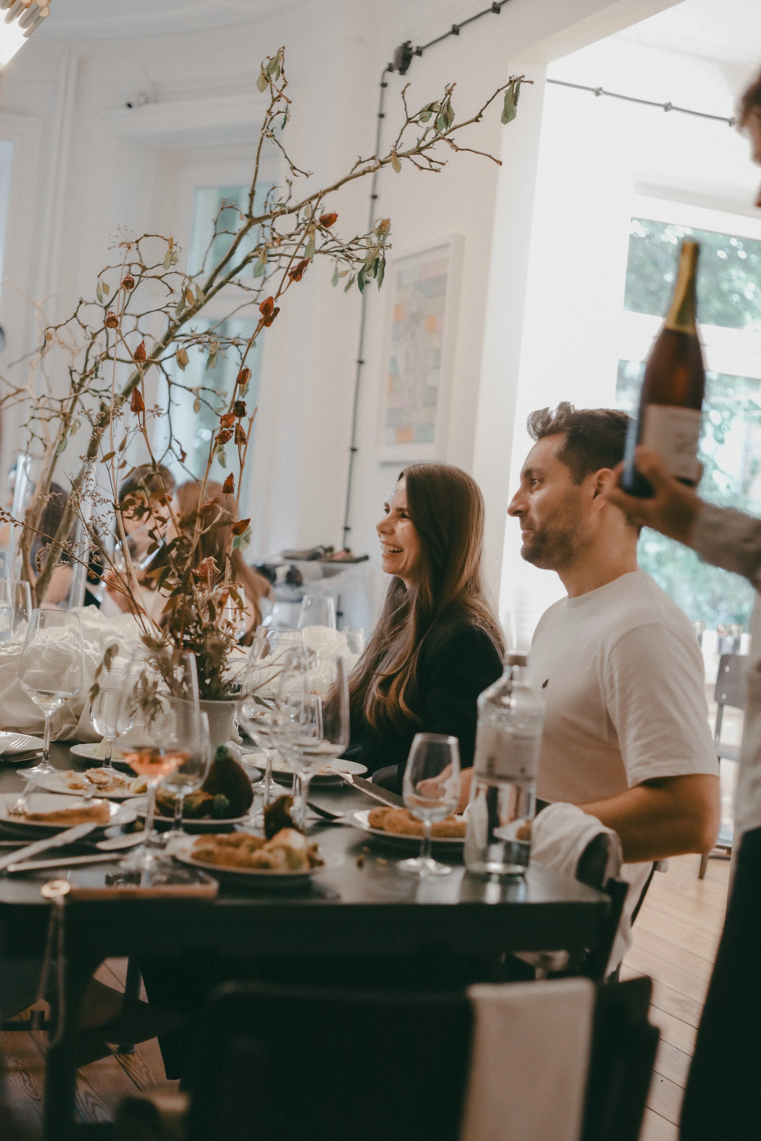 Menschen sitzen bei einem festlichen Essen an einem langen Tisch mit Wein- und Wassergläsern, Speisen und Dekorationen, im Hintergrund eine große Fensterfront und eine Wand mit Kunstwerken, es scheint ein besonderer Anlass zu sein.