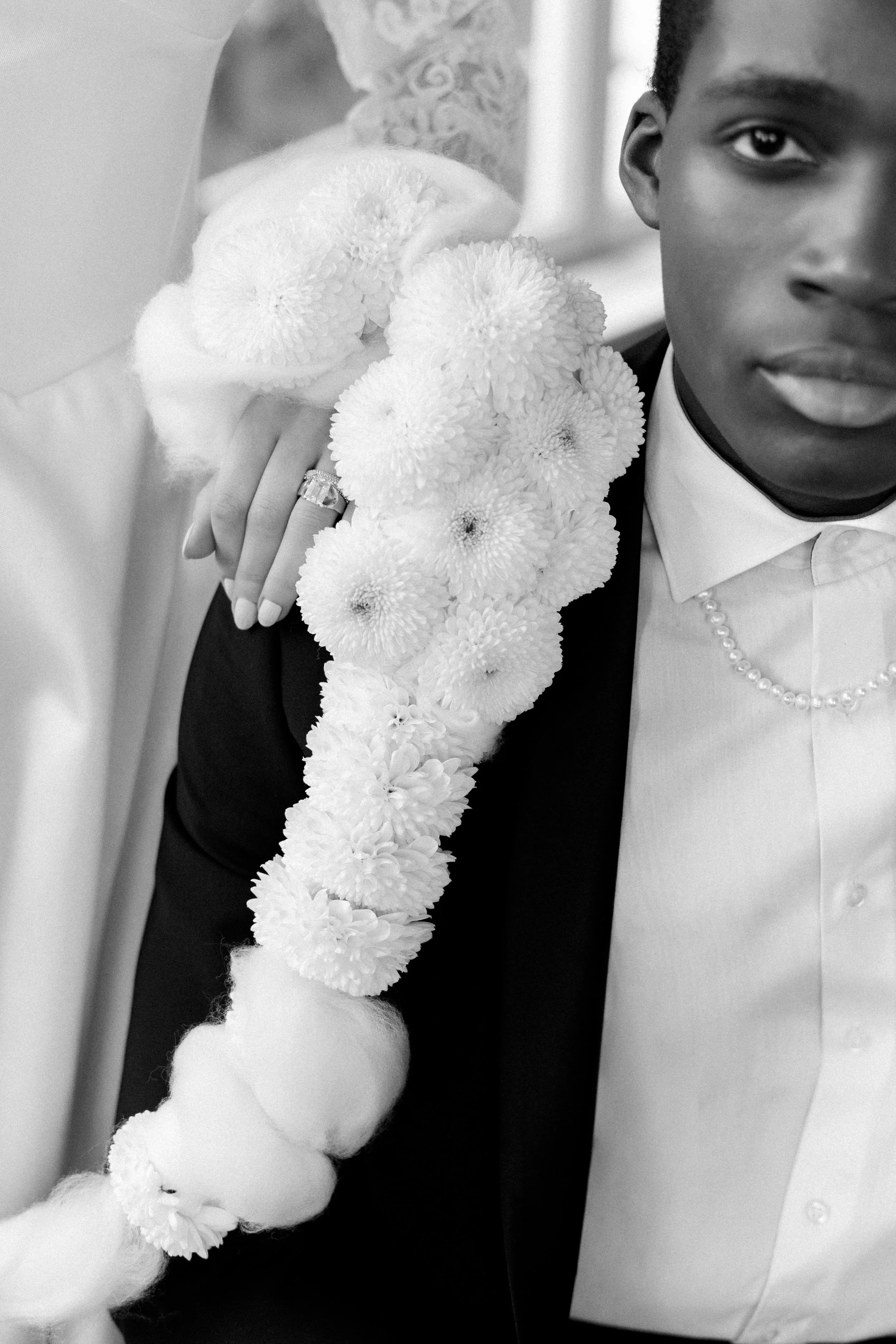 Schwarz-Weiß-Foto eines dunkelhautigen Jungen, der einen weißen Hemd und einen schwarzen Anzug trägt, mit weißen Blumen, die an seiner Schulter hängen.