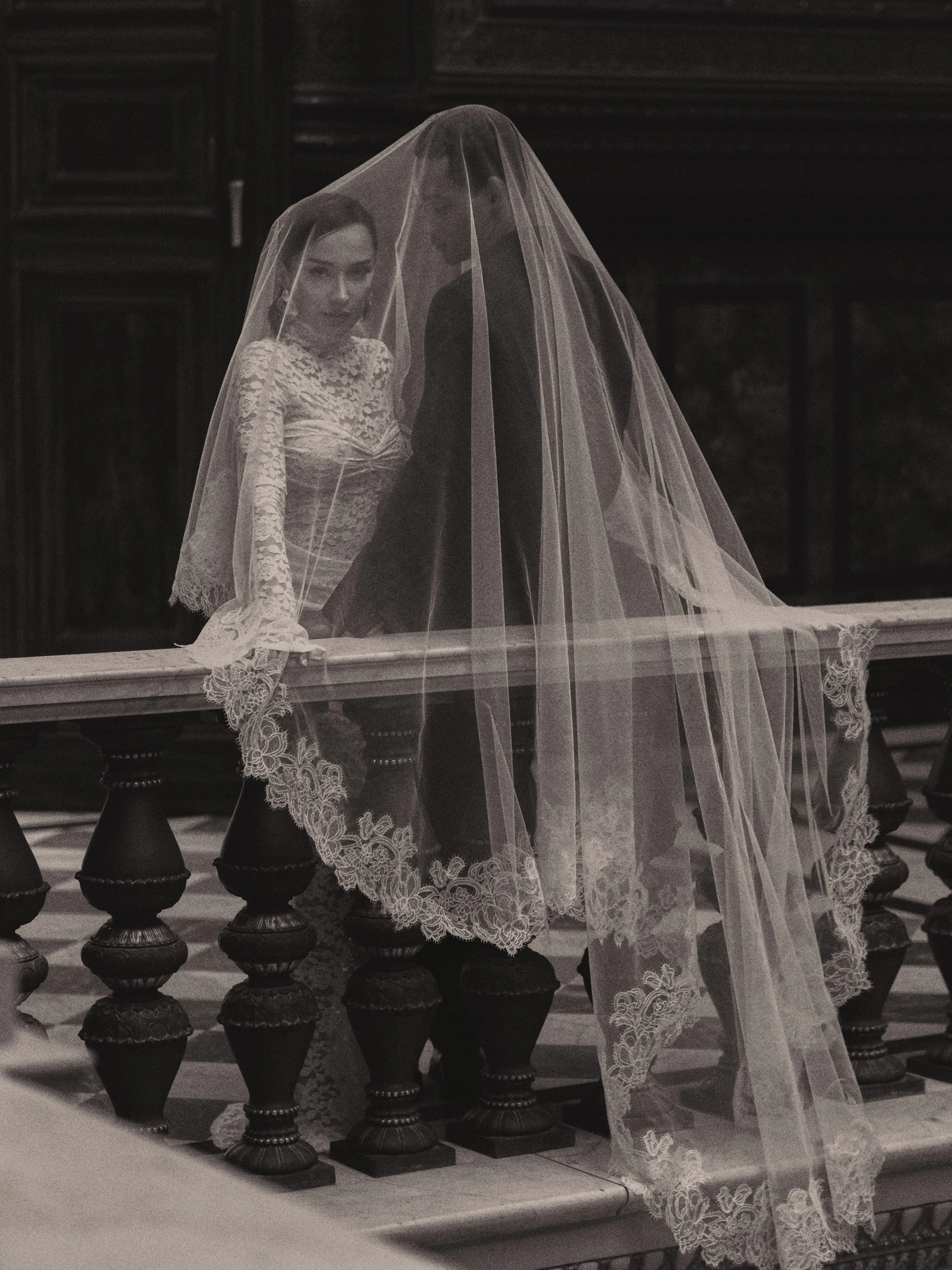 Eine Frau in einem weißen Spitzenkleid und Schleier sitzt an einem Schachbrett. Der Hintergrund ist dunkel und die Szene wirkt dramatisch und elegant.