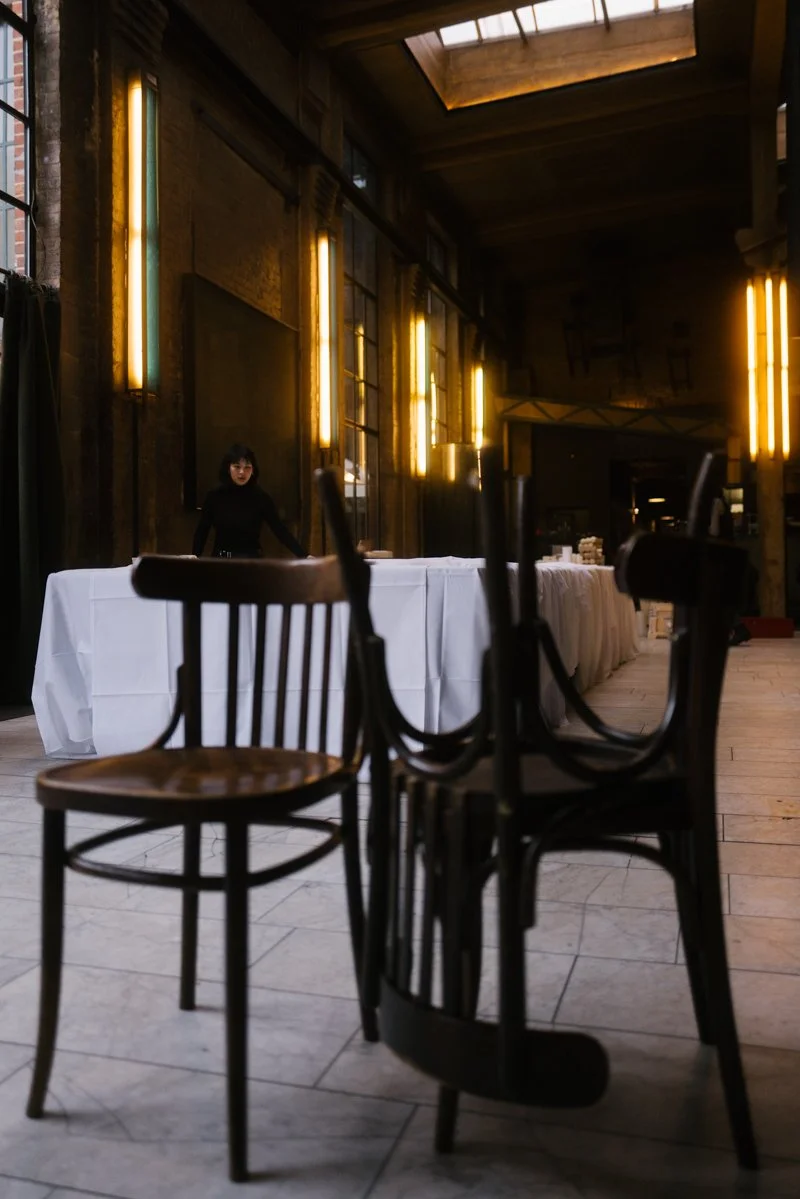 Ein leerer Raum mit schwarzen Stühlen, weißen Tischen und warmem gelbem Licht, möglicherweise eine Veranstaltungs- oder Restaurantlocation.