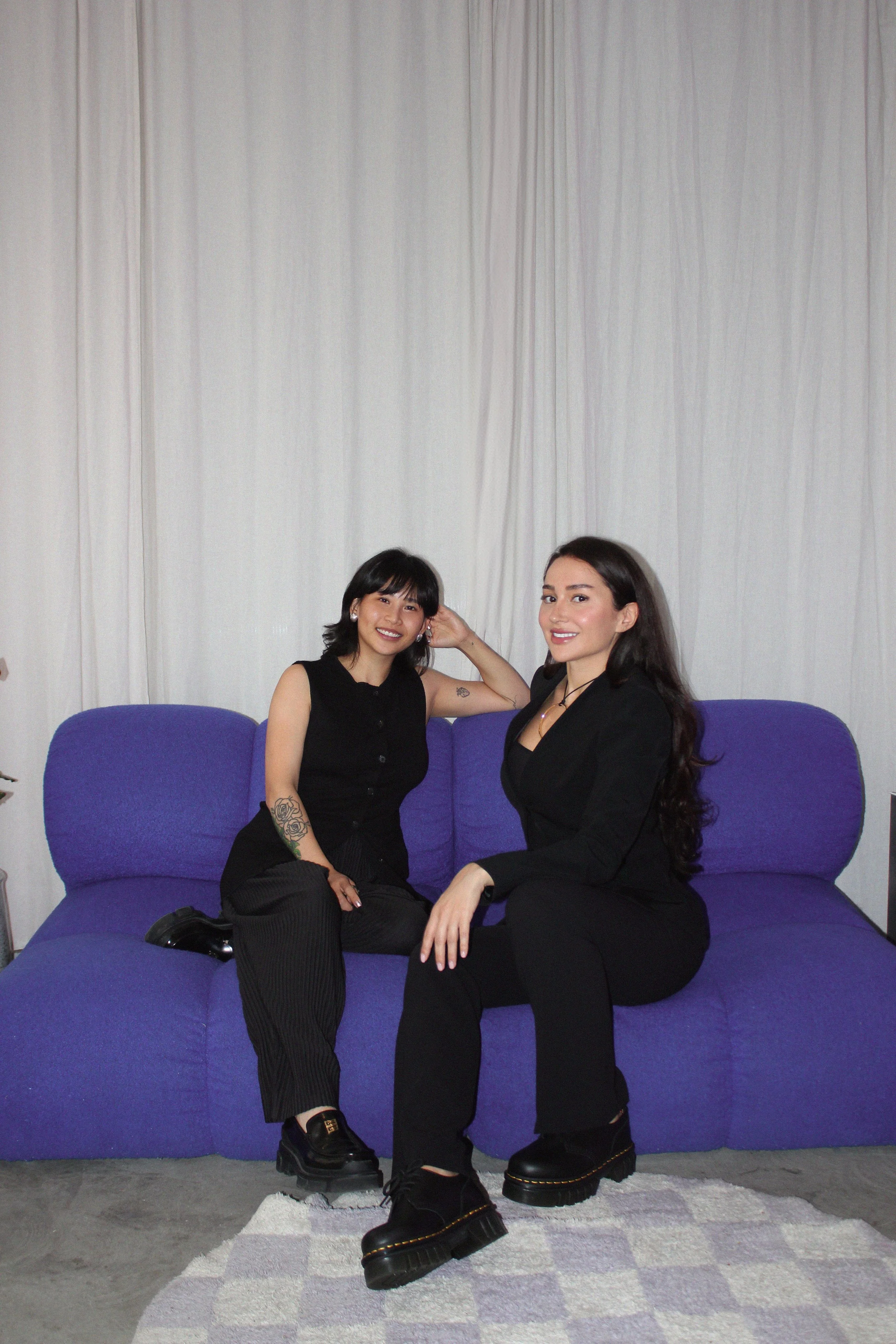 Two women sitting on a purple sofa in a room with light-colored curtains; one is wearing a black sleeveless top and pants, the other is wearing a black blazer and pants.