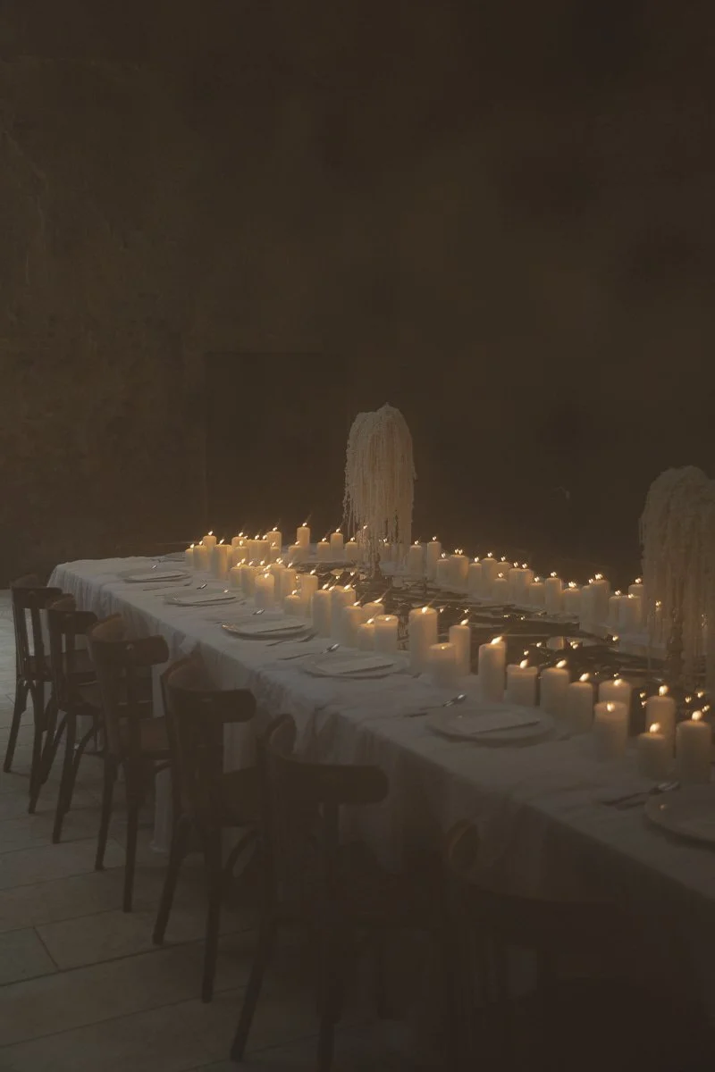 Ein länglicher Esstisch mit weißen Tellern, Besteck und vielen Kerzen in einem dunklen Raum mit steinernen Wänden, dekoriert für eine elegante Veranstaltung.