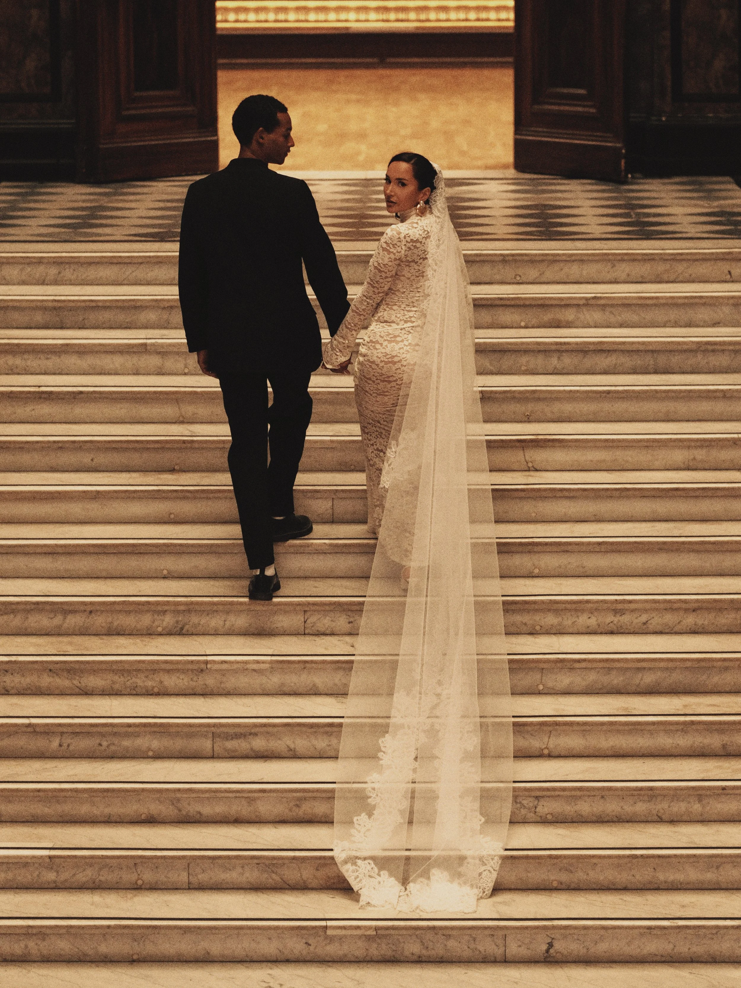 Ein Brautpaar auf Treppen in einem eleganten Innenraum, die Braut trägt ein langes, elegantes Hochzeitskleid mit Schleier, der Bräutigam trägt einen schwarzen Anzug.