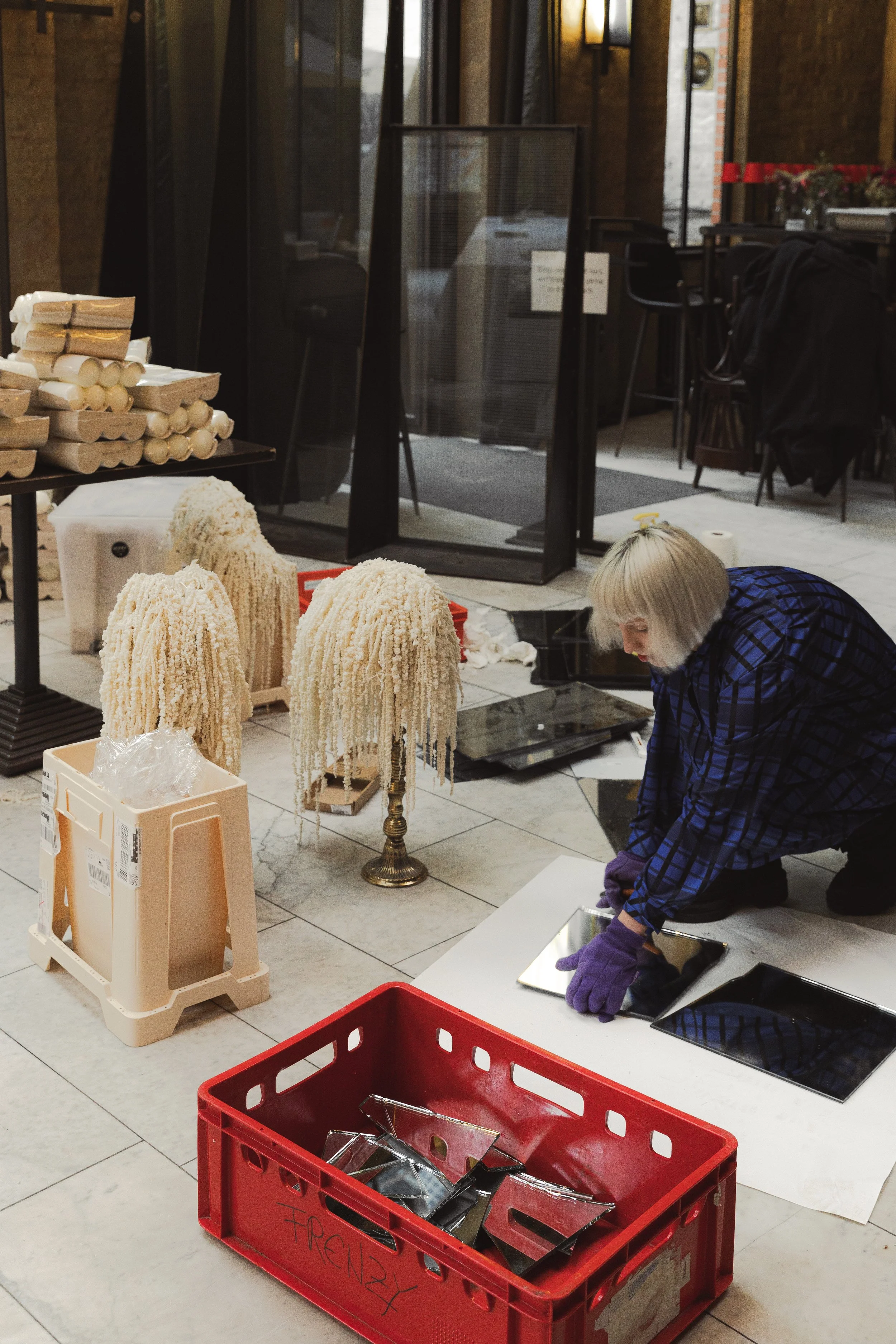 Eine Person mit blonder Kurzhaarfrisur und lila Handschuhen arbeitet an einem DIY-Projekt auf dem Boden eines Innenraums. Im Vordergrund steht eine rote Kiste mit mehreren Metallteilen. Auf einem Tisch sind kreative Skulpturen aus textilen Materialie