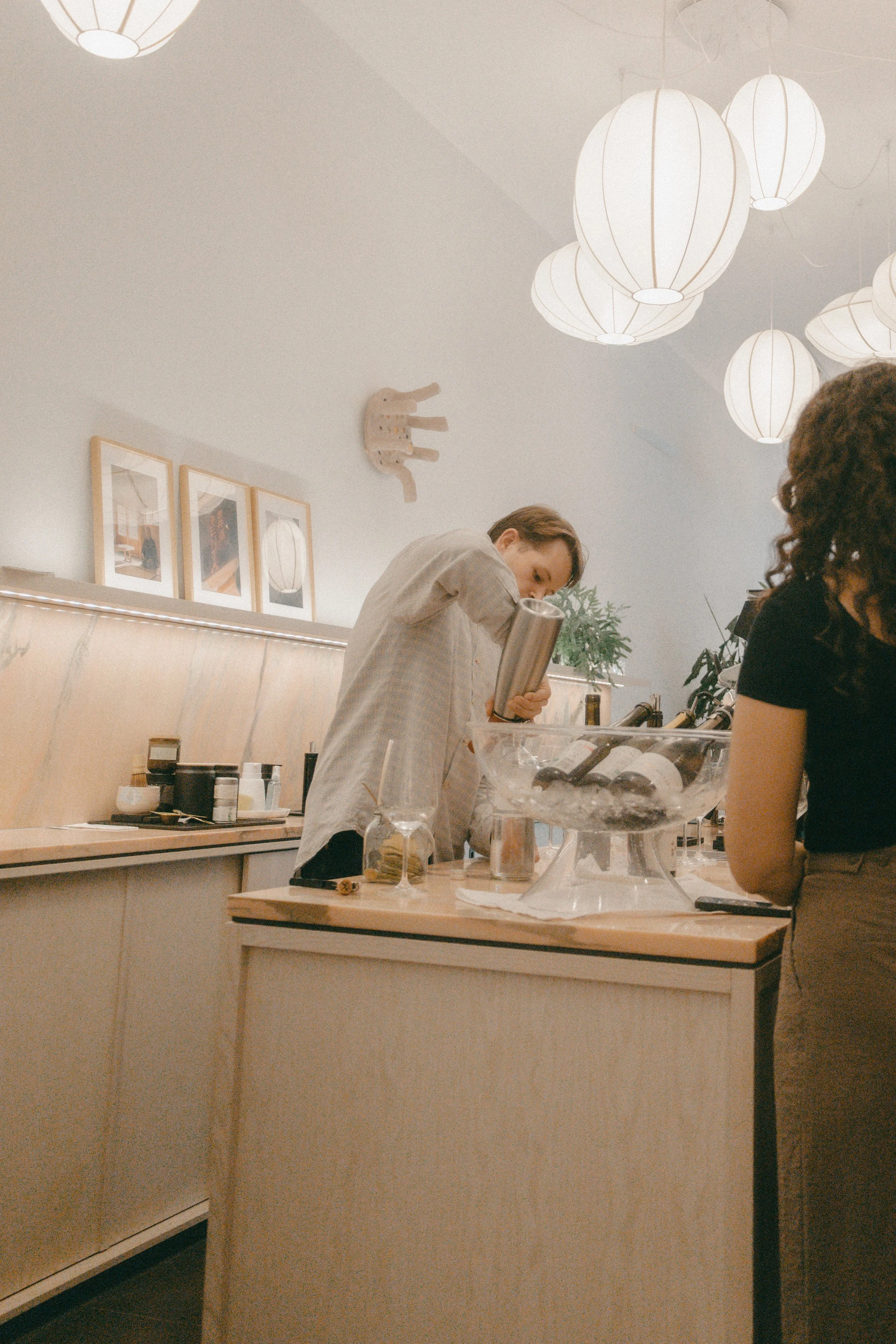 Innenraum einer Bar oder eines Restaurants mit drei weißen, papierähnlichen Lampions an der Decke, mehreren Bildern an der Wand, und zwei Menschen, die an einer Bar arbeiten und Geschirr spülen.