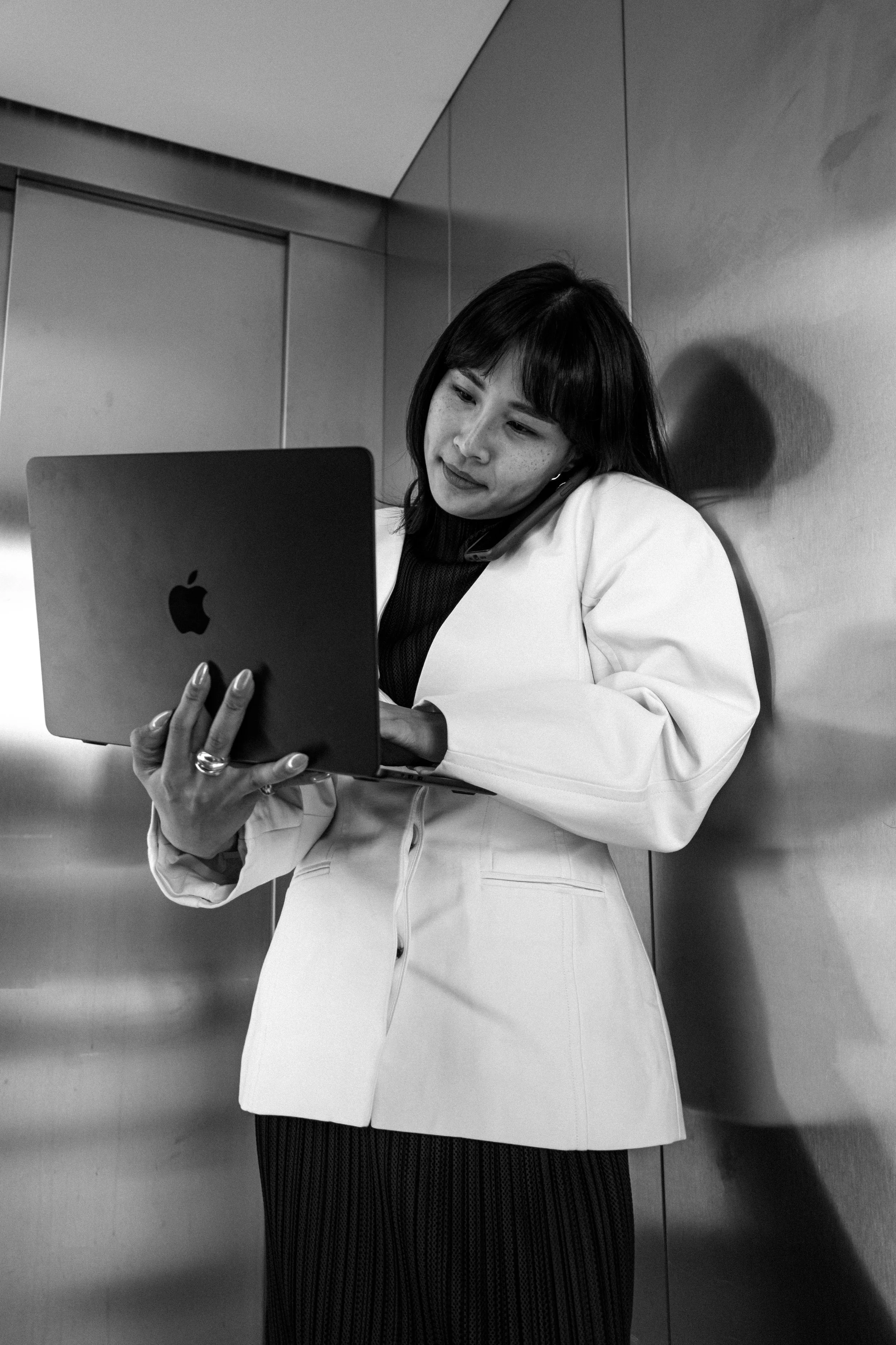 Schwarz-weiß-Foto einer jungen Frau in einem weißen Blazer, die in einem Aufzug mit einem iPad in der Hand arbeitet und auf ihren Telefonanruf hört.