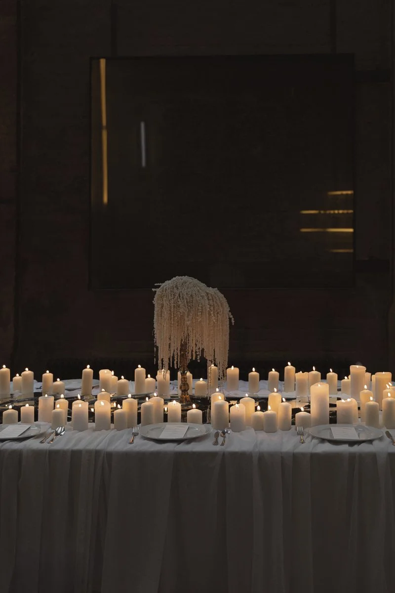 Ein festlich gedeckter Tisch mit vielen Kerzen, einer eleganten Blumenarrangement in der Mitte, weißen Tellern, Besteck und weißen Servietten, in einem dunklen Raum mit Kerzenschein.