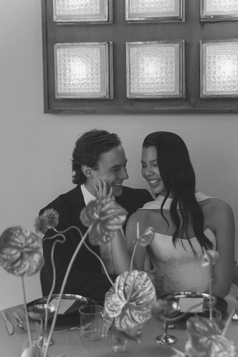 Zwei Menschen bei einer Feier, die sitzen an einem Tisch, lachen und scheinen sich zu umarmen, mit einem Blumenstrauß im Vordergrund und einem Wandbild im Hintergrund.