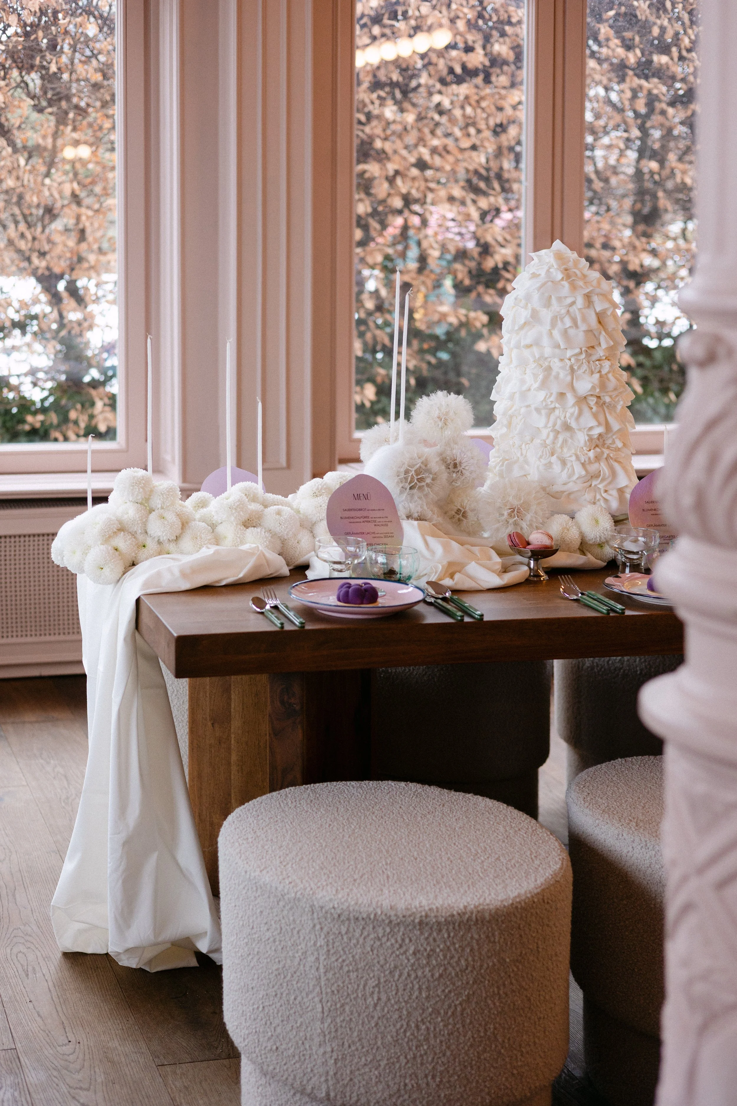 Ein gedeckter Tisch mit weißen Blumen, Kerzen und Dessert, im Hintergrund große Fenster mit Blick auf herbstlich gefärbte Bäume.