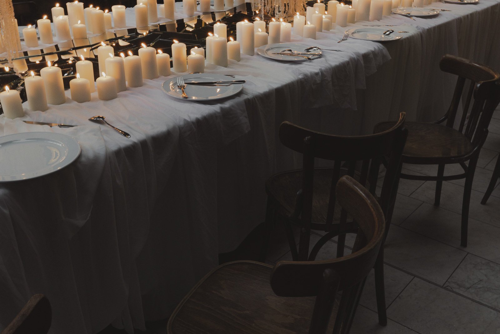 Ein rechteckiger Speisetisch mit weißen Kerzen und leeren Tellern, Besteck liegt auf den Tellern, dunkle Holzmöbel, wahrscheinlich für eine festliche Abendveranstaltung oder Dinner