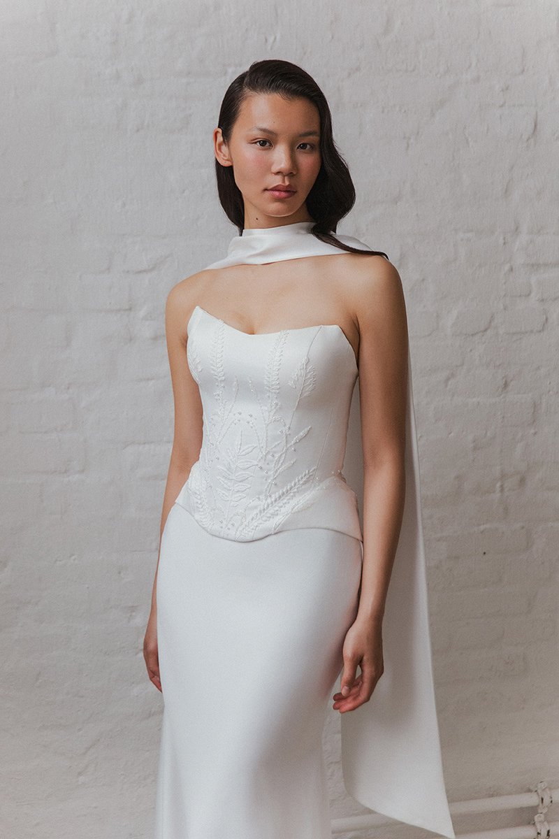 Eine Frau trägt ein elegantes weißes Kleid mit passender Halskette vor einer weißen Ziegelwand.