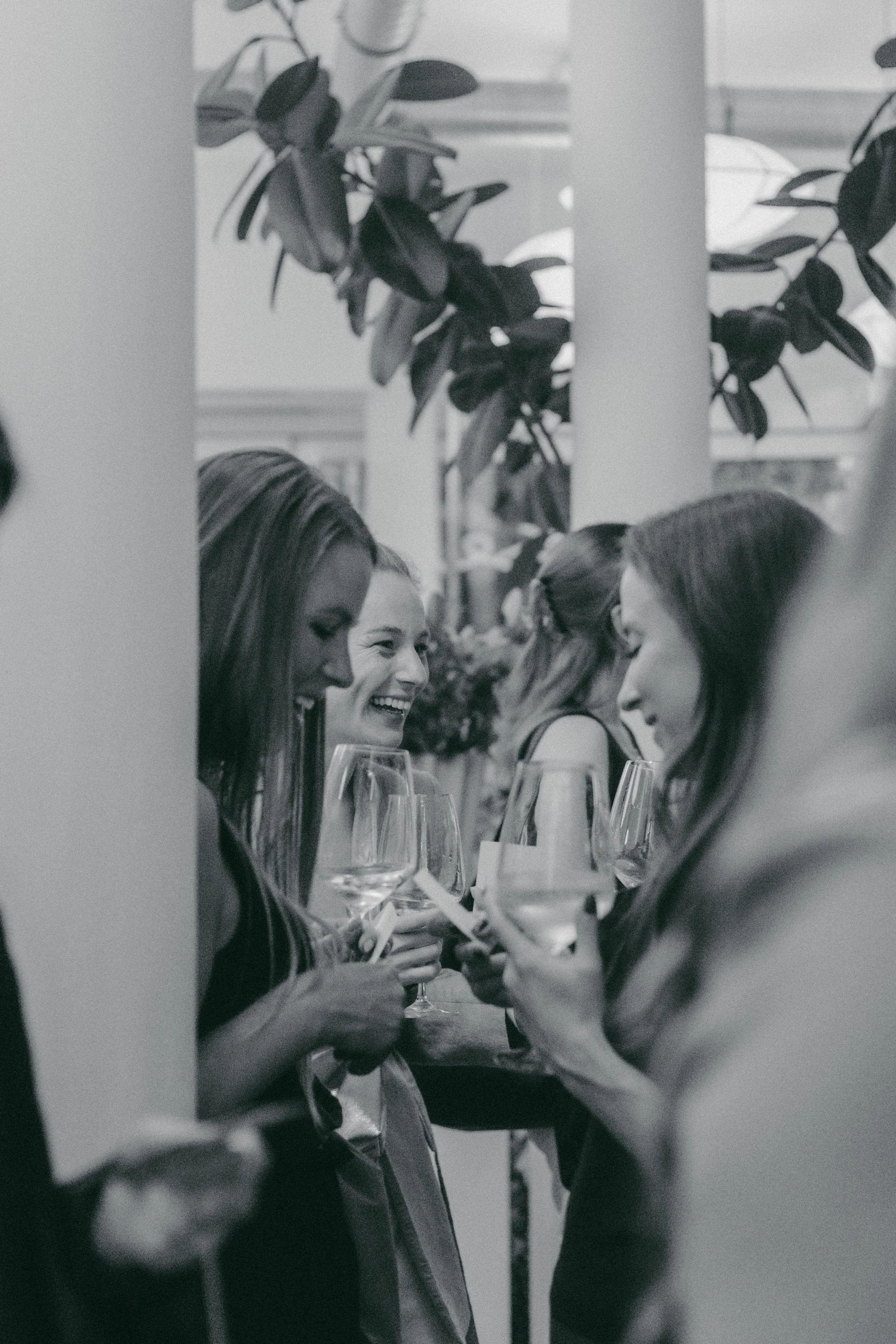 Drei Frauen sitzen an einem Tisch, unterhalten sich und trinken Wein, im Hintergrund sind Pflanzen zu sehen.