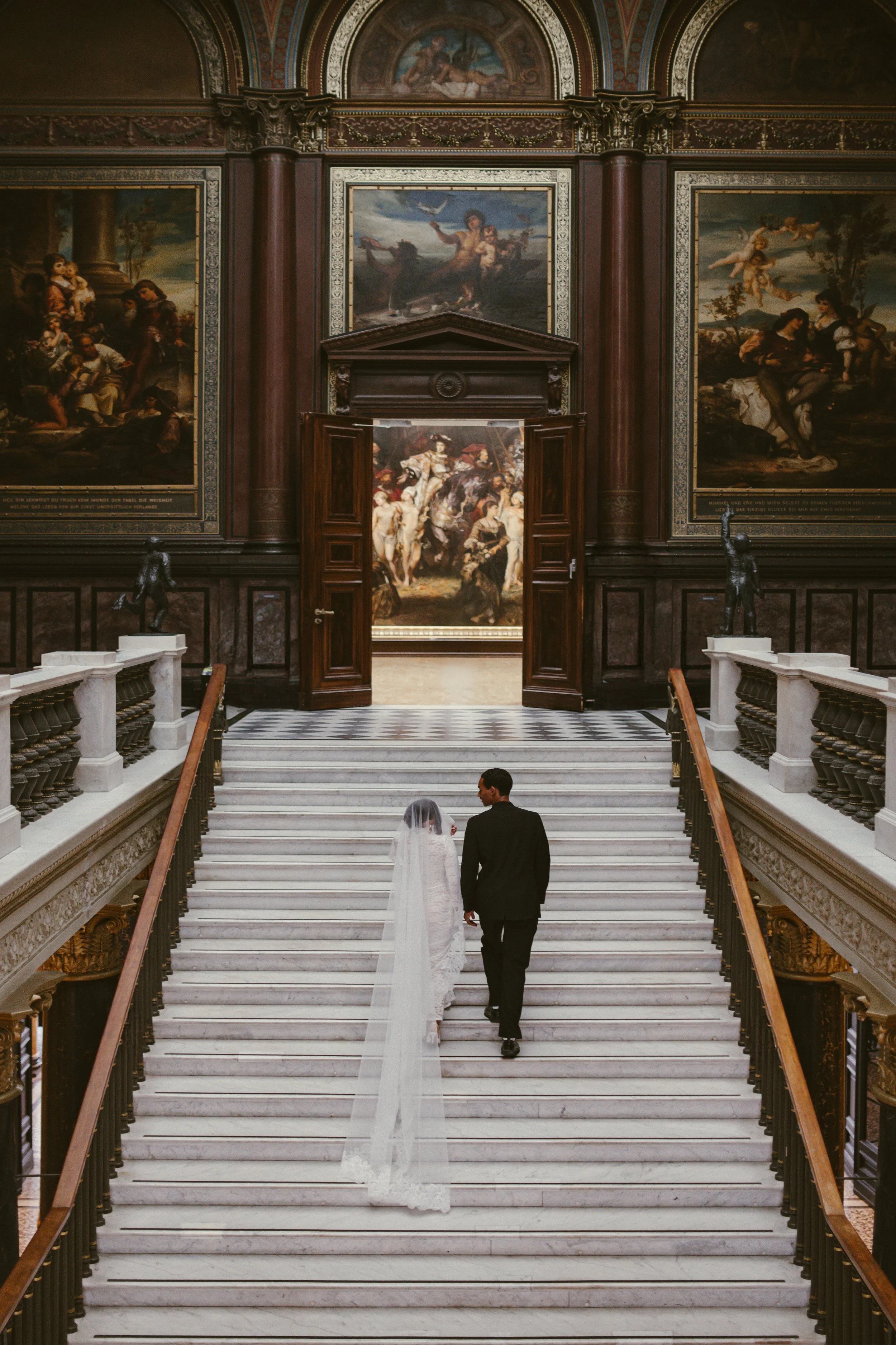 Ein Hochzeitspaar, das die Treppe in einem prunkvollen, historischen Gebäude hinauffährt, mit zahlreichen Gemälden und Verzierungen an den Wänden