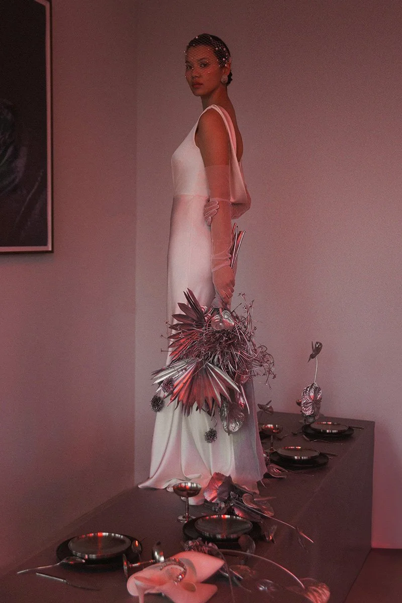 Eine Frau in einem weißen Abendkleid steht auf einem Tisch, umgeben von silbernen Geschirr und Dekorationsstücken, die eine elegante Atmosphäre schaffen.