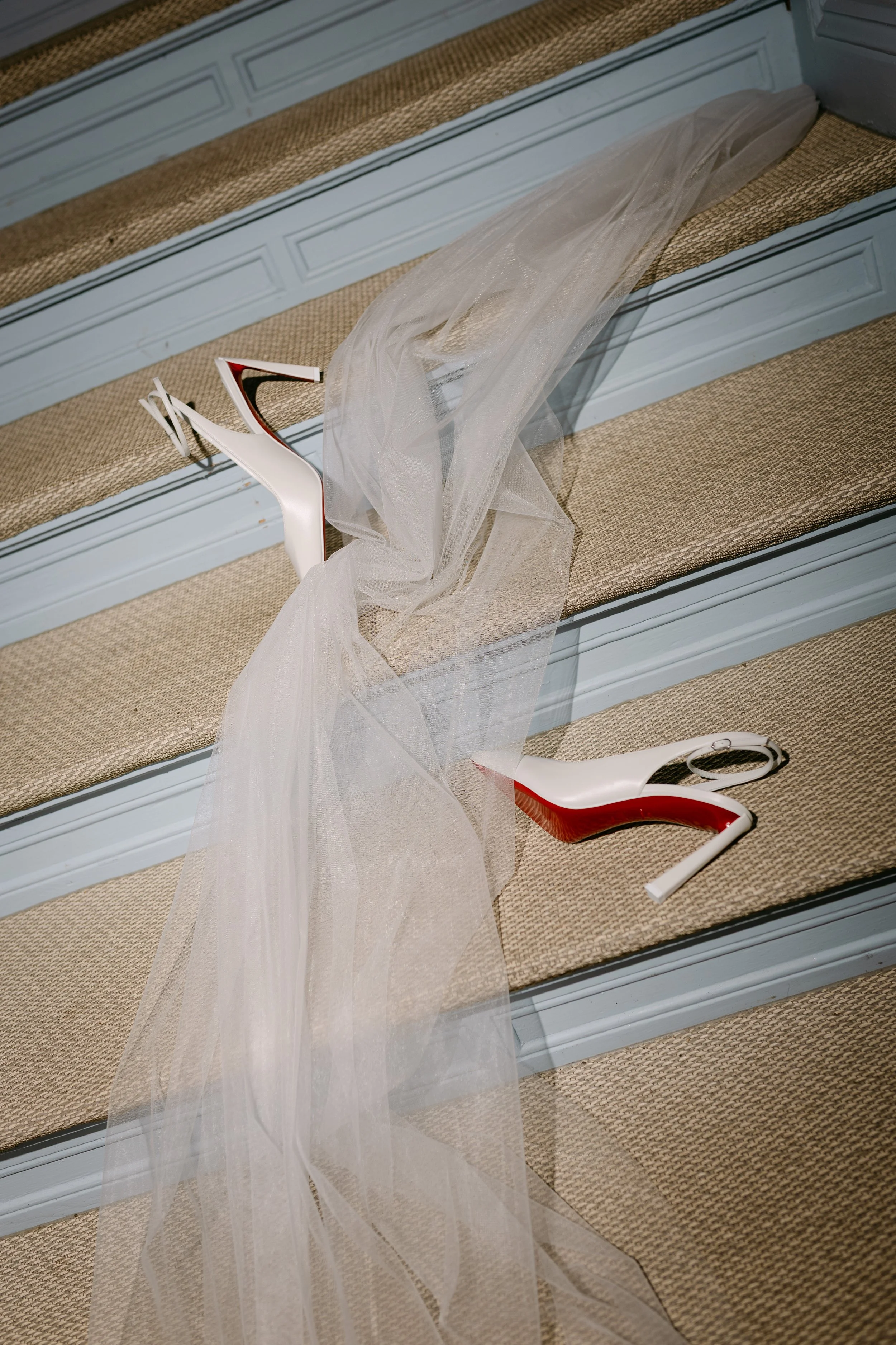 Ein weißer hoher Schuh mit rotem Absatz liegt auf Treppenstufen, eine weiße Tüll- oder Organza-Band liegt auf den Stufen.