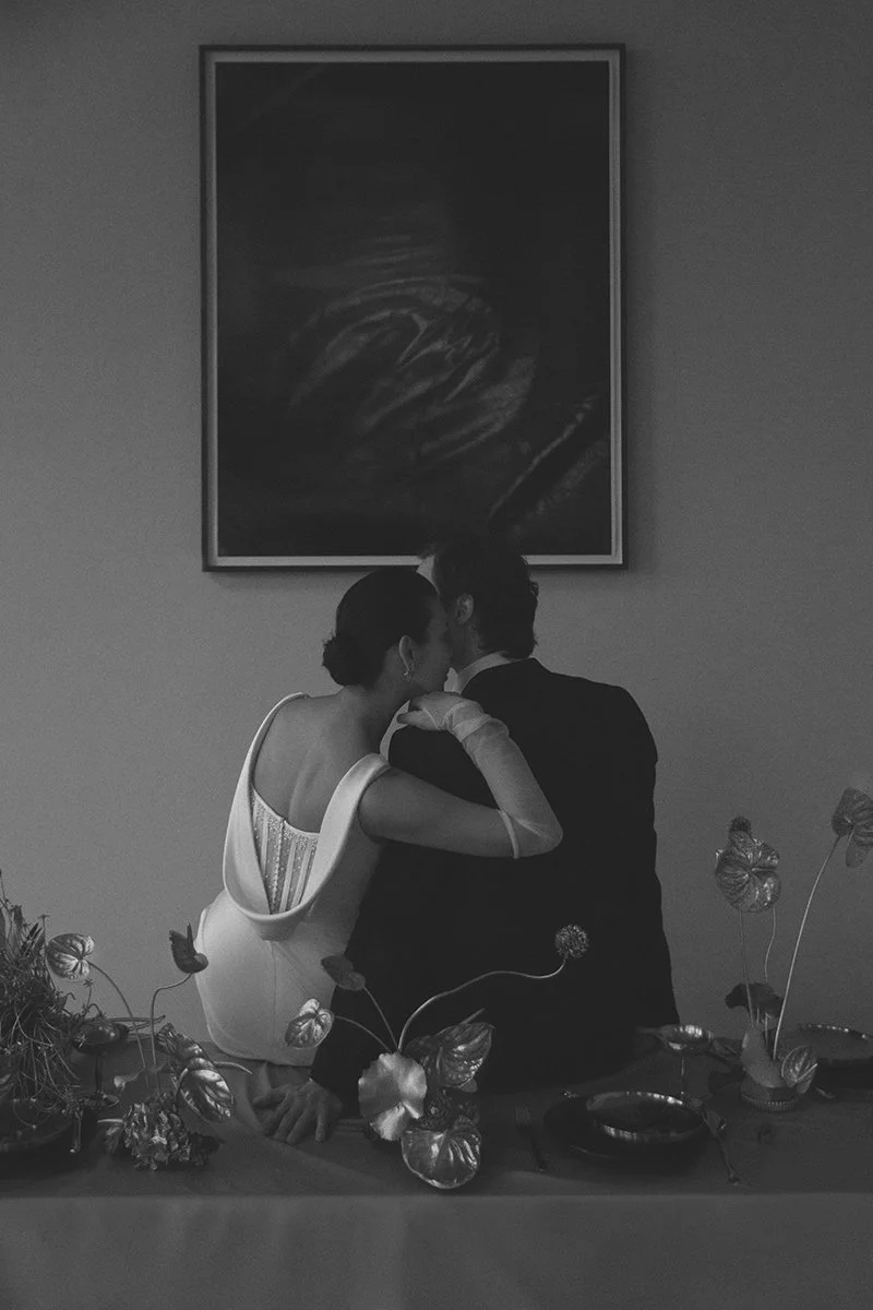 Ein schwarzes und weißes Bild eines Paares, das sich küsst, mit Blumen im Vordergrund und einem Gemälde an der Wand im Hintergrund.
