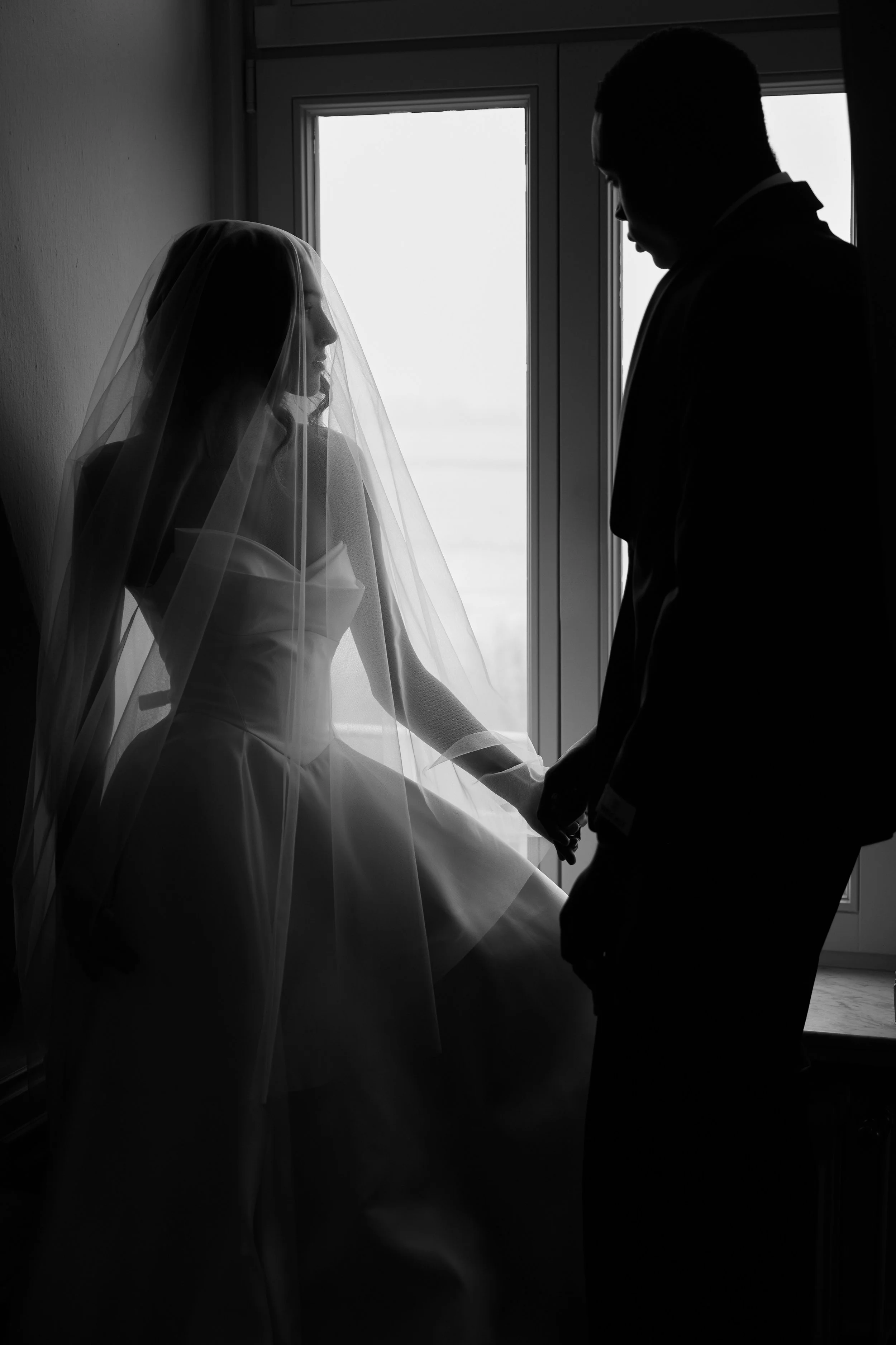 Schwarz-weiß-Fotografie eines Paares, das sich hält, im Hintergrund ein Fenster mit natürlichem Licht, das eine Silhouette erzeugt.