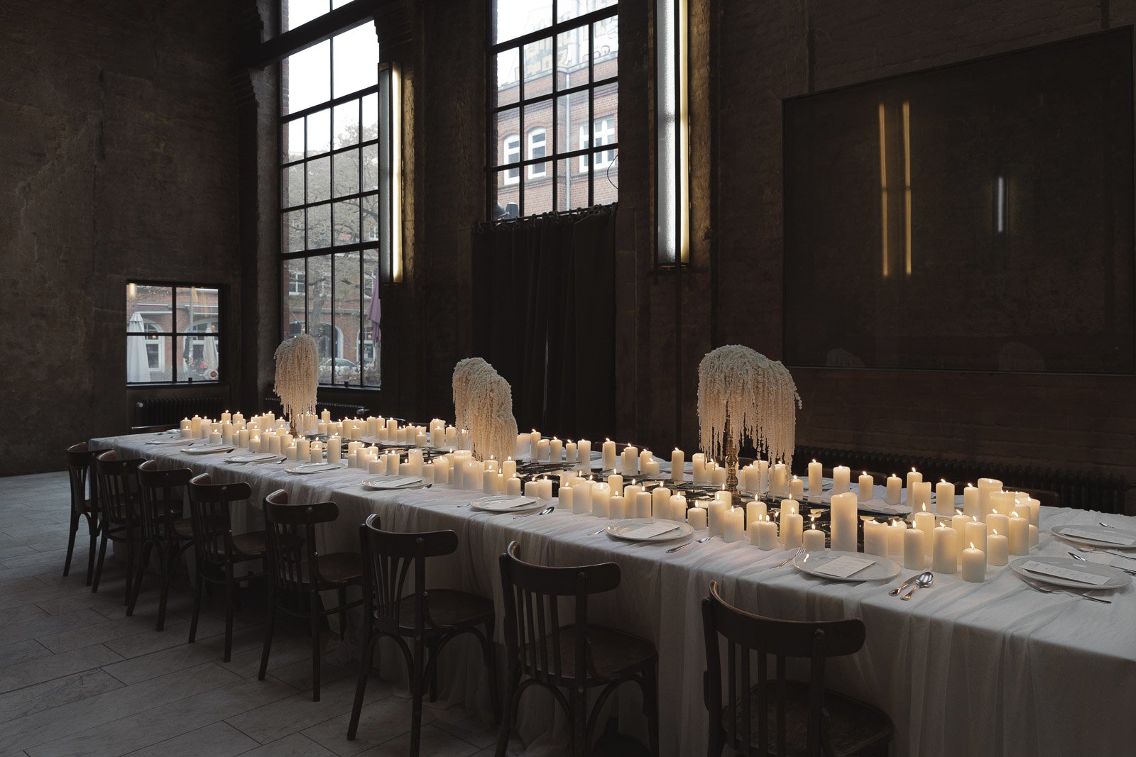 Ein langer Festtisch mit weißen Kerzen, die für eine Dekoration bei einer Veranstaltung sorgen. Der Tisch ist mit weißen Tellern, Besteck und Papieren gedeckt, und es gibt hohe weiße Blumenarrangements als Dekoration. Das Zimmer hat große Fenster mit