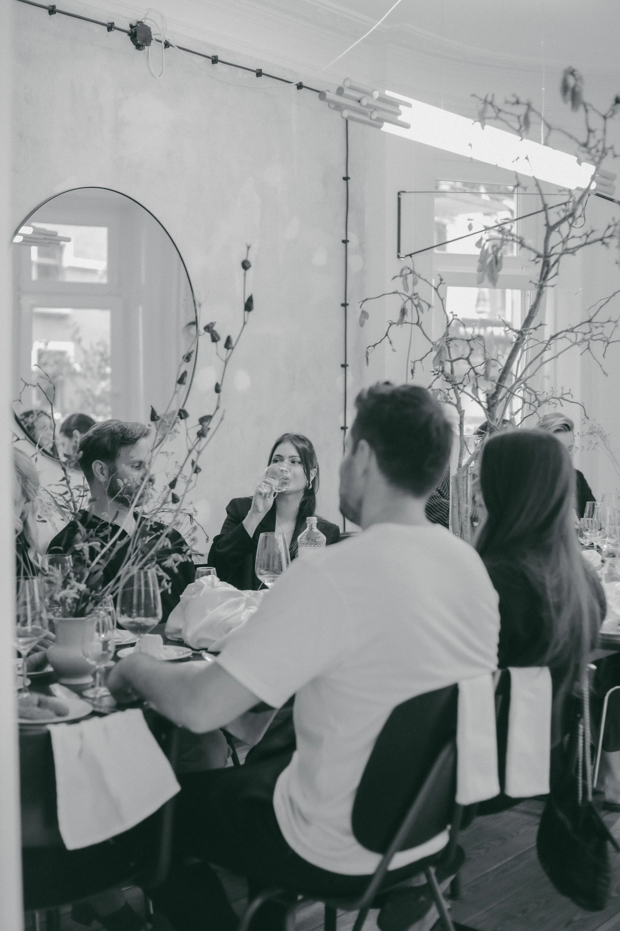 Menschen sitzen an einem Esstisch, trinken und sprechen in einem wohlgehüteten Restaurant, der Raum ist hell erleuchtet mit natürlichen Licht durch die Fenster, Pflanzen und moderne Dekorationen sind sichtbar.