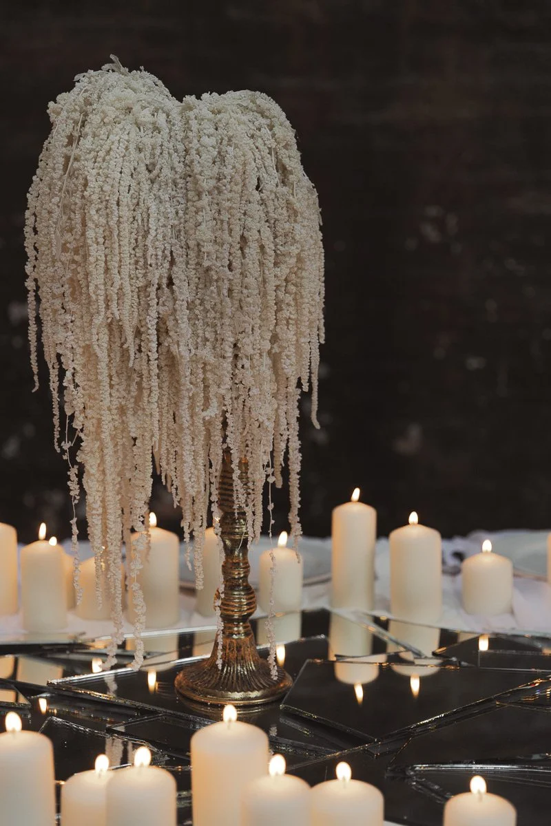 Ein Kerzenständer mit einer dekorativen Anordnung aus weißen Zimtstauden, umgeben von brennenden weißen Kerzen auf einem Spiegel, auf dunklem Hintergrund.