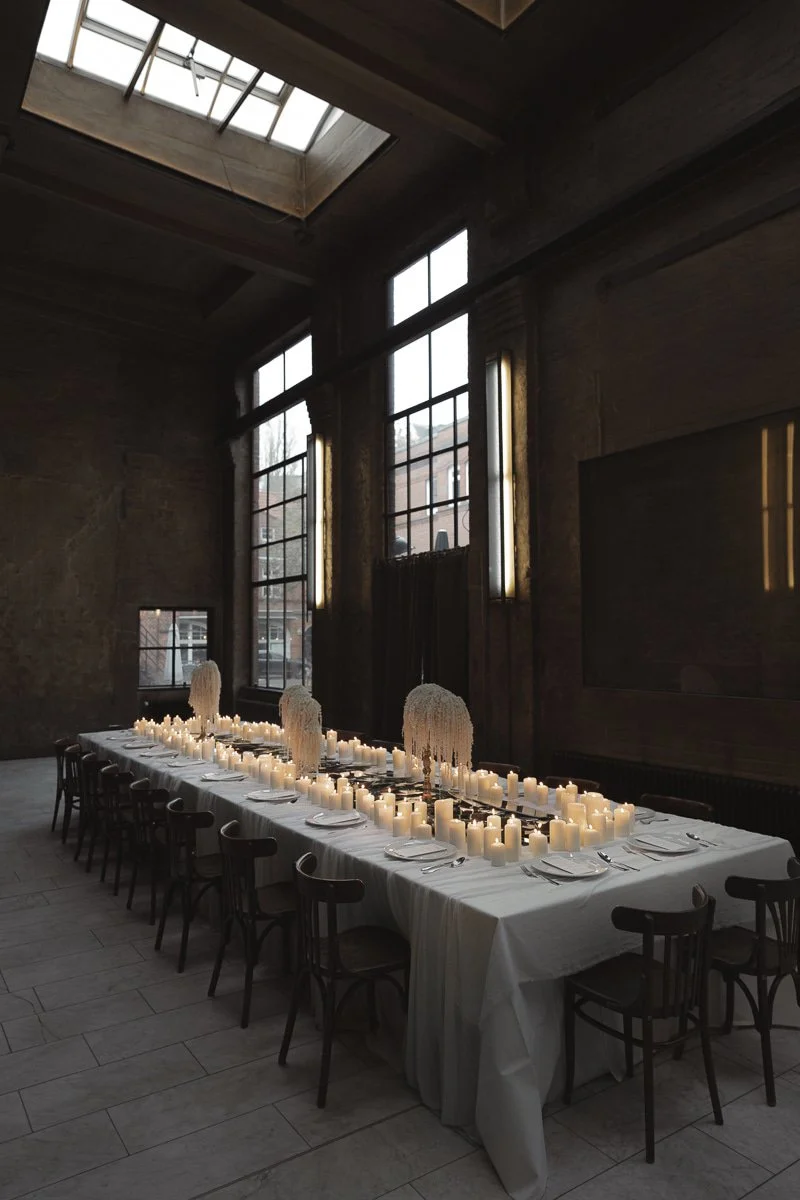 Ein langer Esstisch mit weißer Tischdecke, Kerzen und Dekorationen in einem dunklen, industrialartigen Raum mit hohen Fenstern und einem großen Oberlicht.