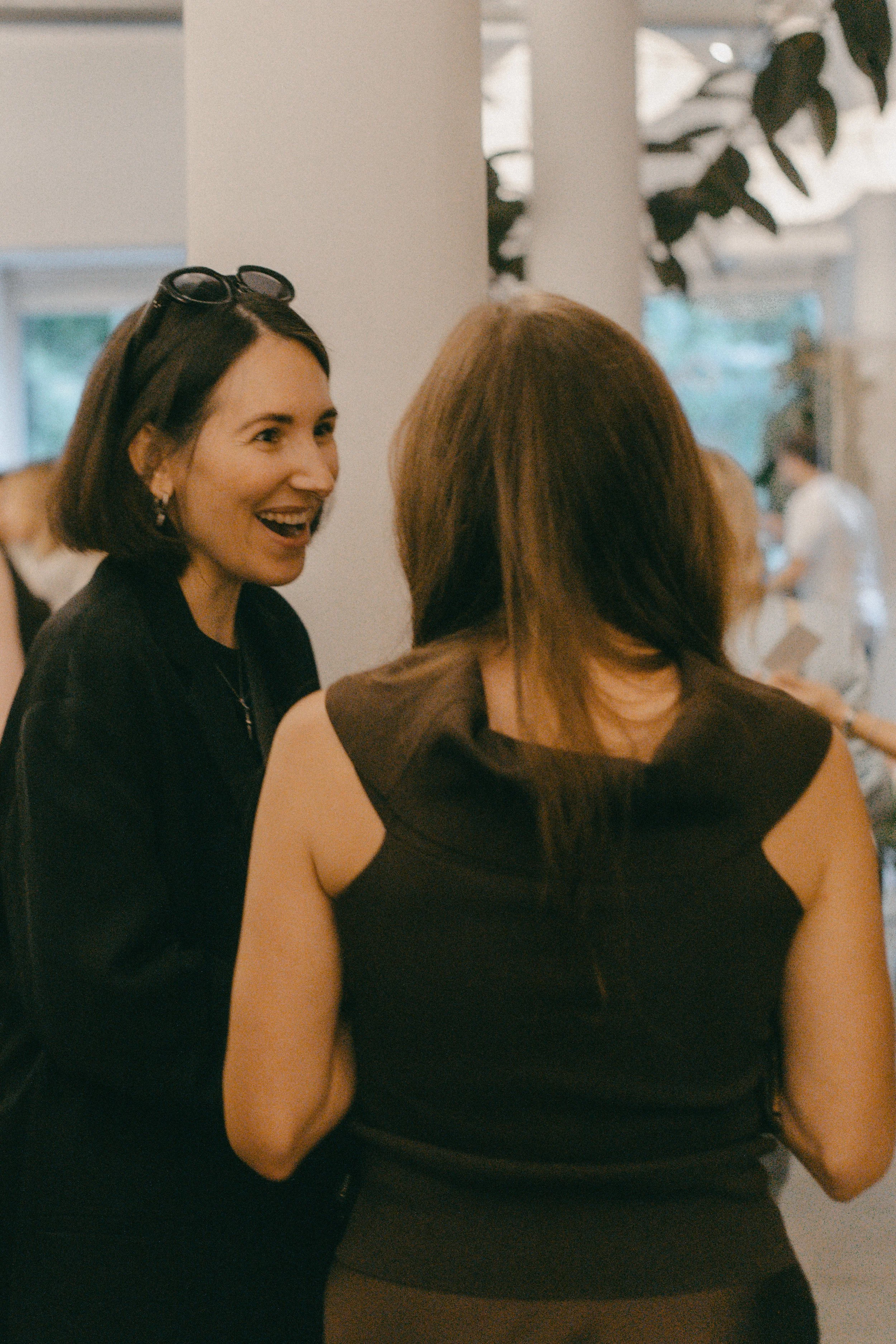 Zwei Frauen im Gespräch bei einer gesellschaftlichen Veranstaltung in einem modernen Raum mit natürlichem Licht und Pflanzen im Hintergrund.