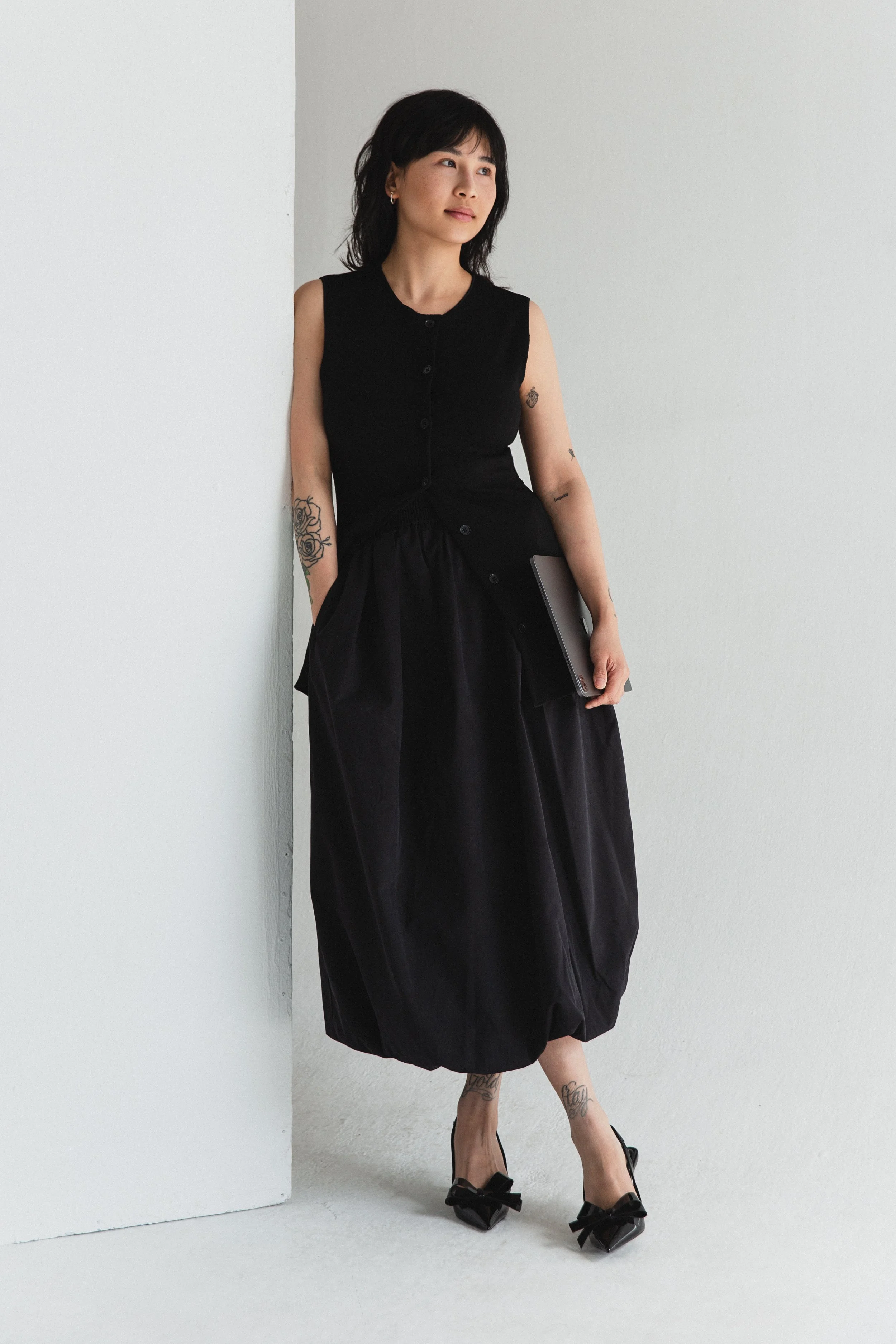 Eine Frau in einem schwarzen Kleid und High Heels steht an einer weißen Wand im Raum, hält einen Laptop in der Hand, hat Tattoos an den Armen und Beinen, kurze schwarze Haare und schaut nach oben.