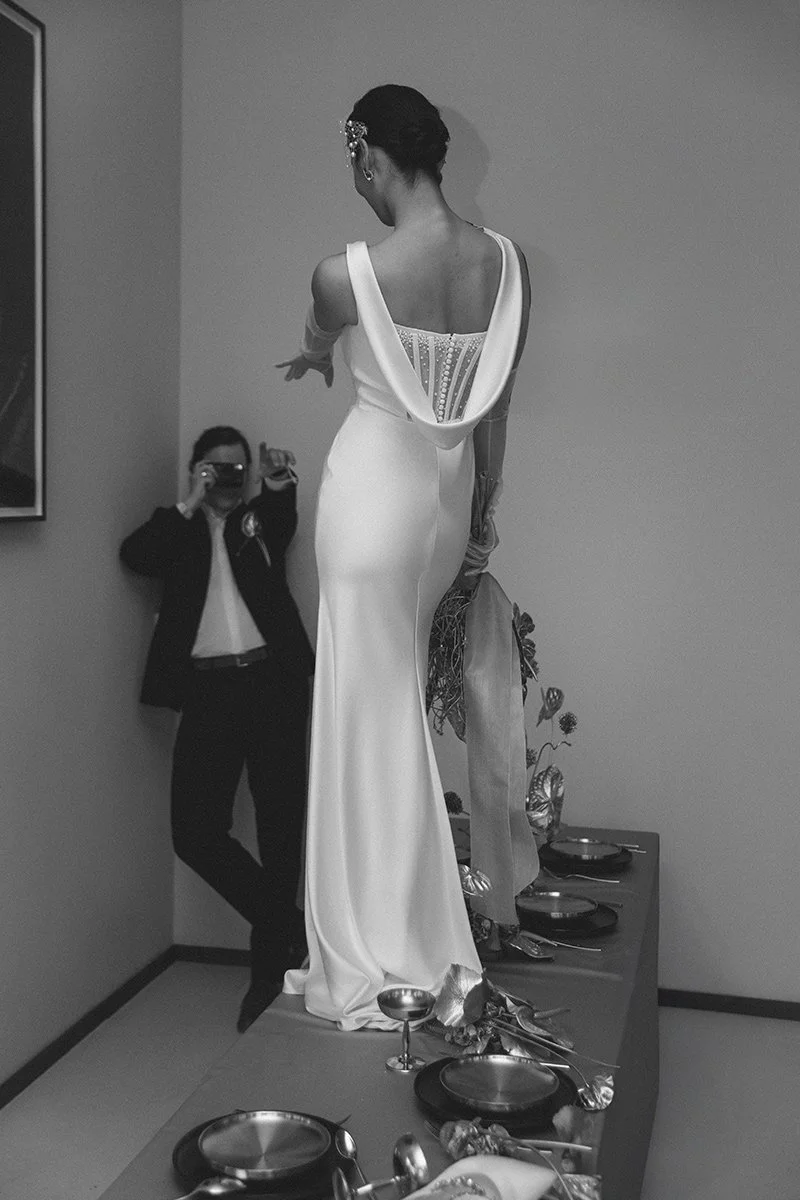 Ein weiteres Modell im weißen Kleid steht auf einem Tisch mit mehreren leeren Tellern und Besteck, während eine Person im Hintergrund Fotos macht.