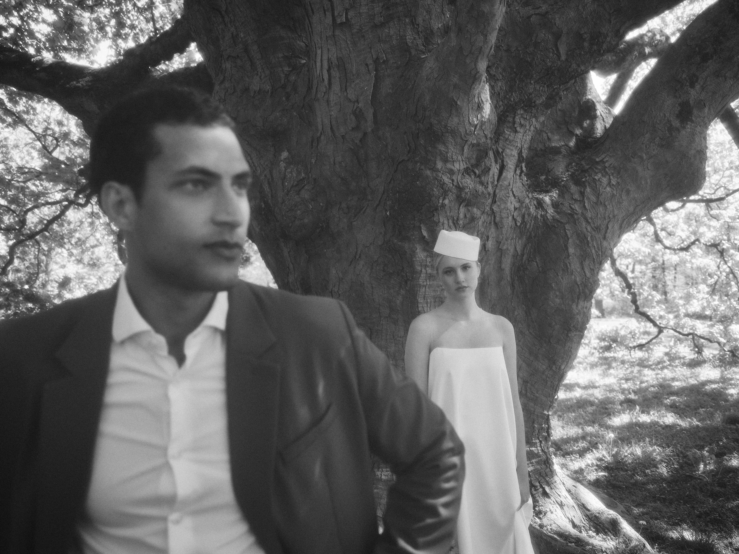 Schwarz-weiß-Foto von einem Mann in einem Anzug und Krawatte im Vordergrund, im Hintergrund ein Baum mit zwei Frauen, eine trägt ein weißes Kleid und eine trägt ein Kleid mit einer Mütze auf dem Kopf, alle befinden sich im Freien in einem Park