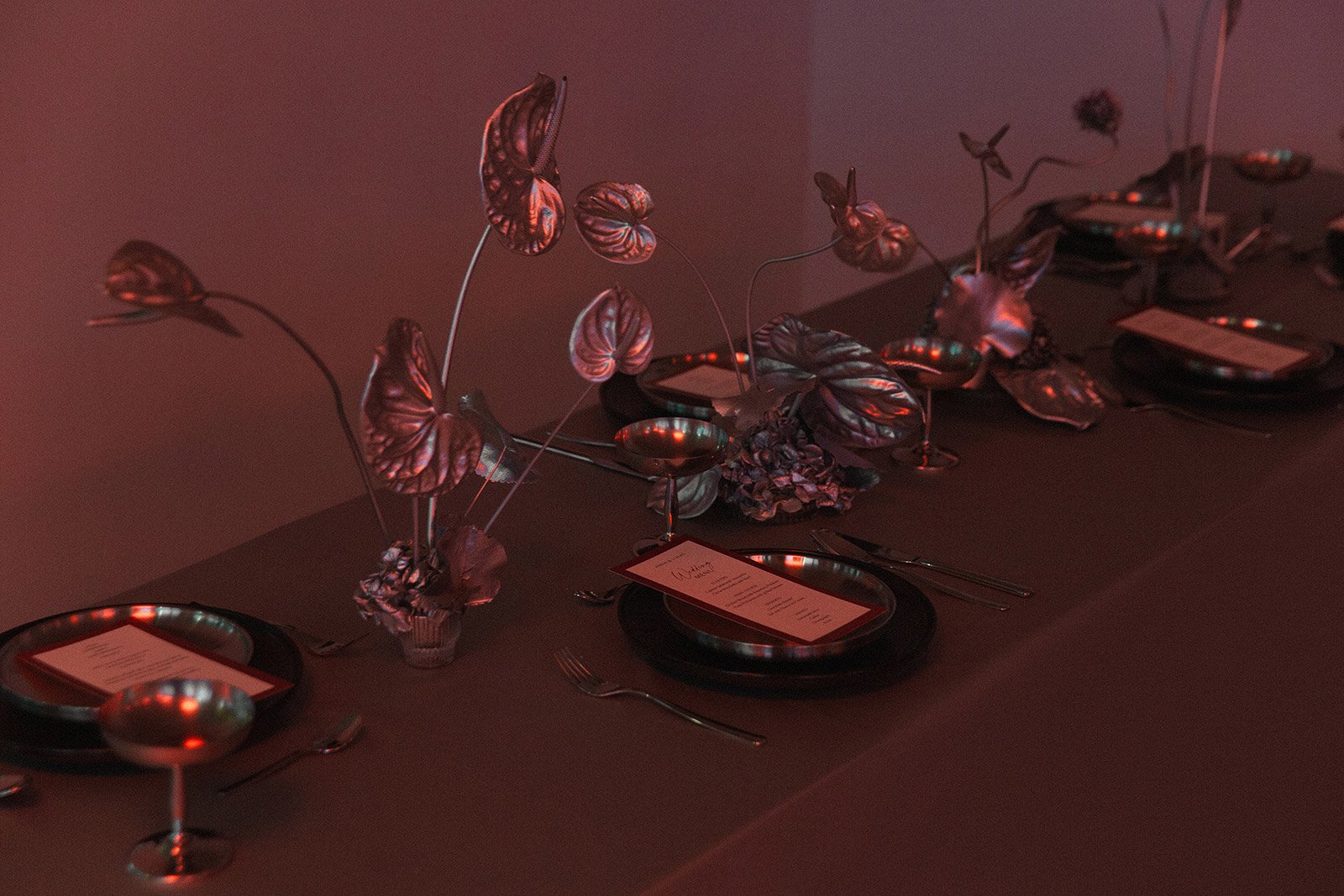 Eleganter gedeckter Tisch mit schwarzen Tellern, silbernen Servietten, silbernen Löffeln, Gabeln und Messern, und einer Dekoration aus metallischen, roségoldfarbenen Blättern und Blumen in kleinen Vasen. Der Raum ist rot beleuchtet.