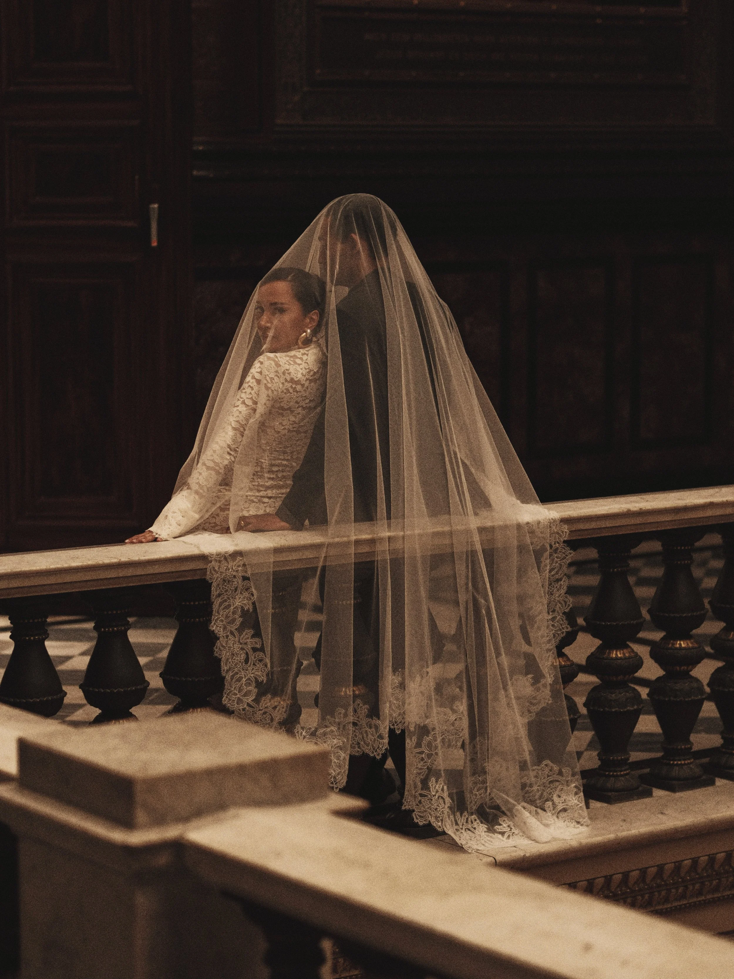 Zwei Frauen in einem Kirchenraum, eine trägt ein Brautkleid mit Spitzenmuster, die andere steht hinter ihr, beide umgeben von dunklem Holzinterieur.
